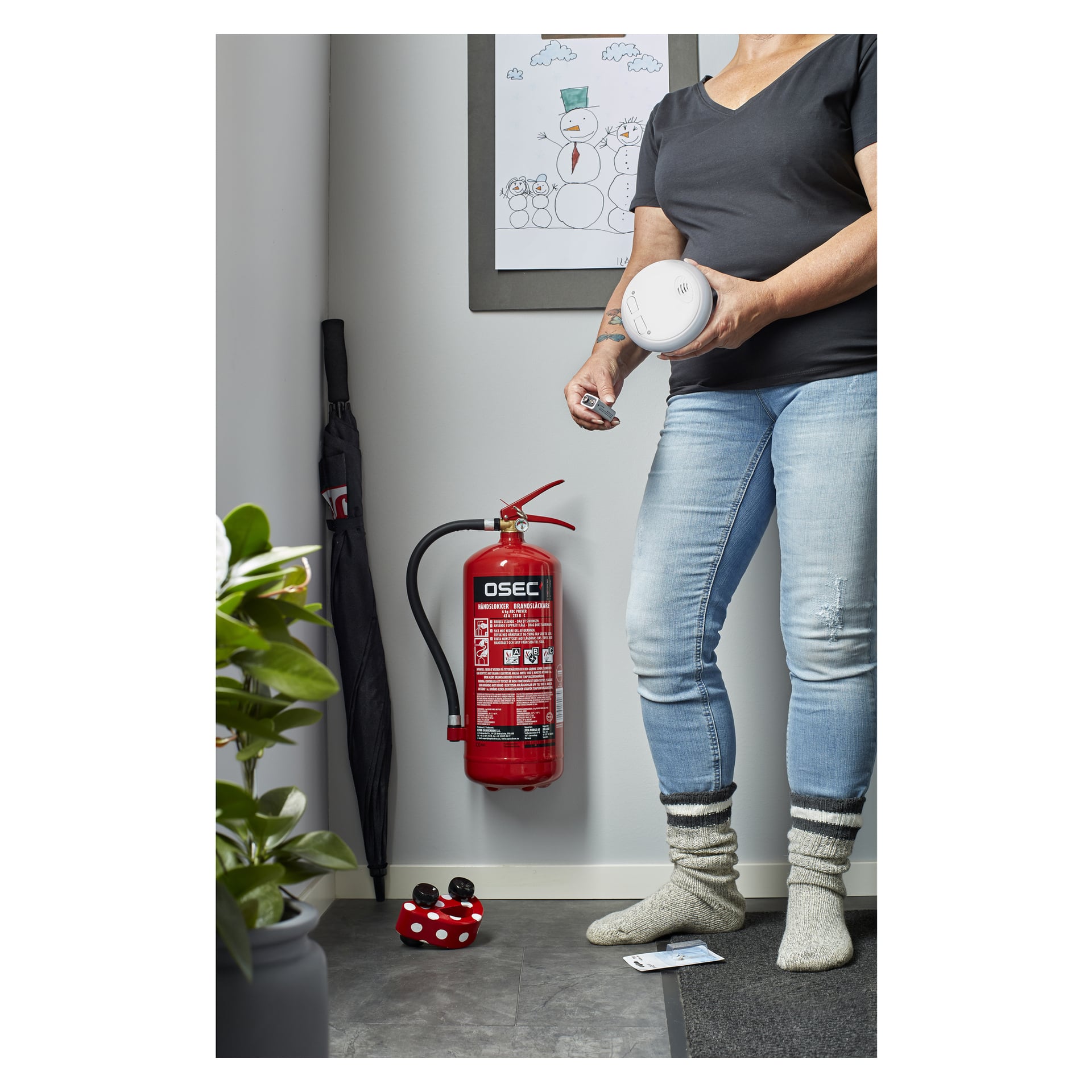 En person med jeans og sokker holder to pakker med Hard Head trådløse røykvarslere ved siden av en rød brannslukker på veggen. En paraply, en plante og et kunstverk med snømenn er i bakgrunnen, og personens ansikt er utenfor bildet.