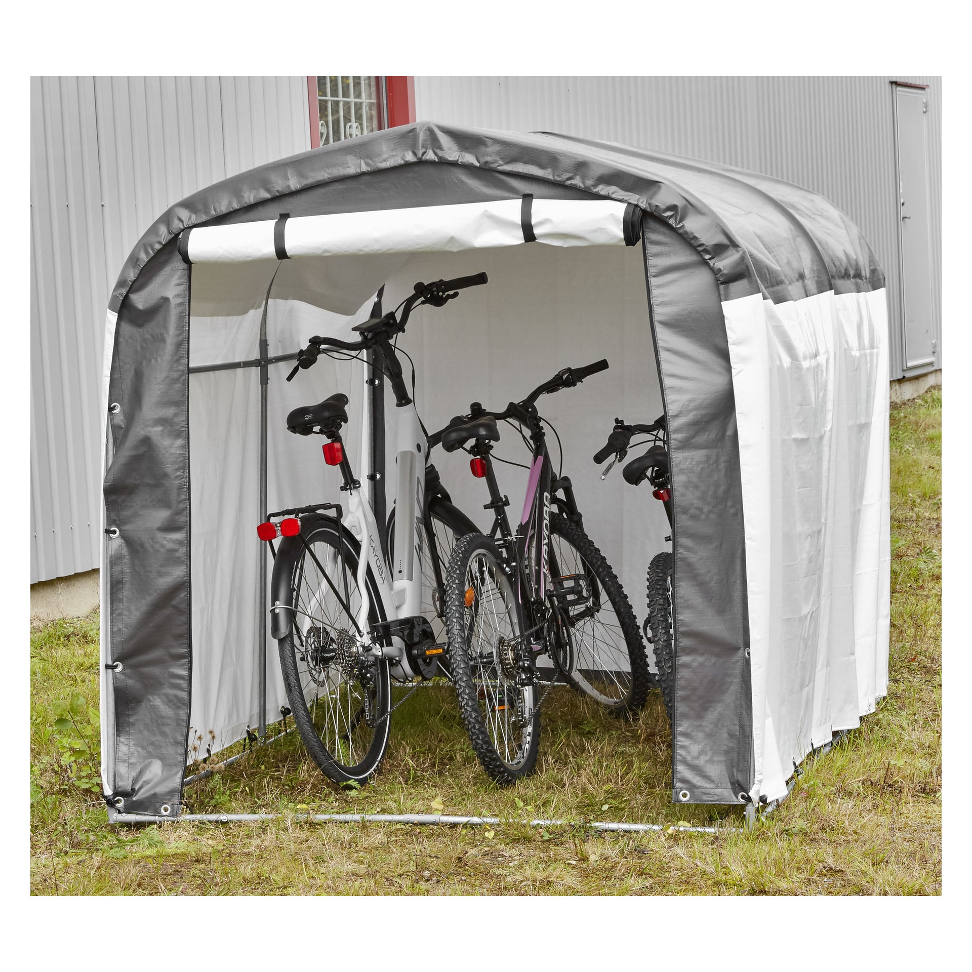 Tre cyklar förvaras i ett tält i Hamron Garage med ett grått och vitt tygskydd, placerat på gräs bredvid en korrugerad metallkonstruktion.