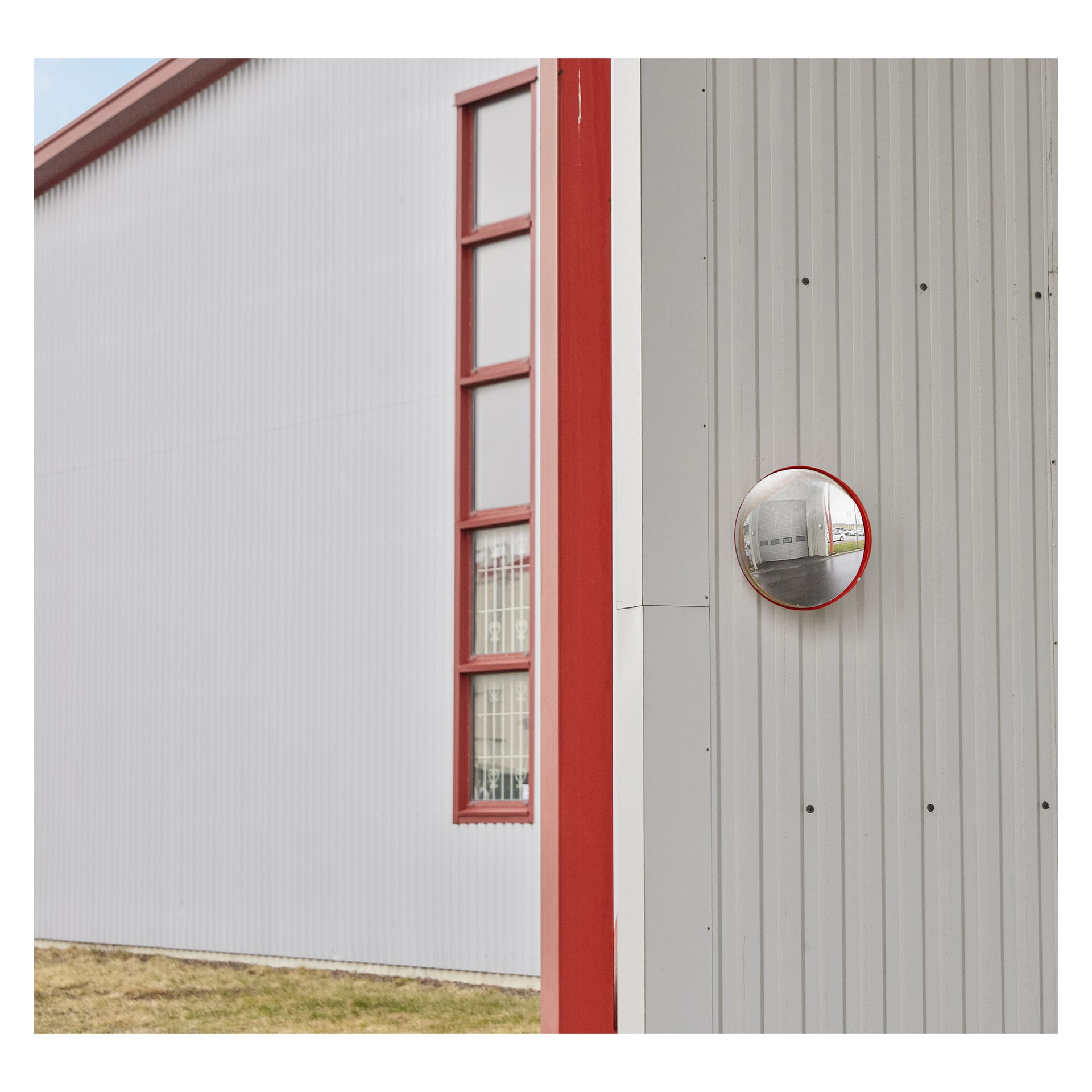 En grå korrugerad metallkonstruktion med röda detaljer har en vertikal rad fönster och en Hamron trafikspegel fäst på väggen, som visar en reflektion av delar av byggnaden. Det växer gräs längs basen.