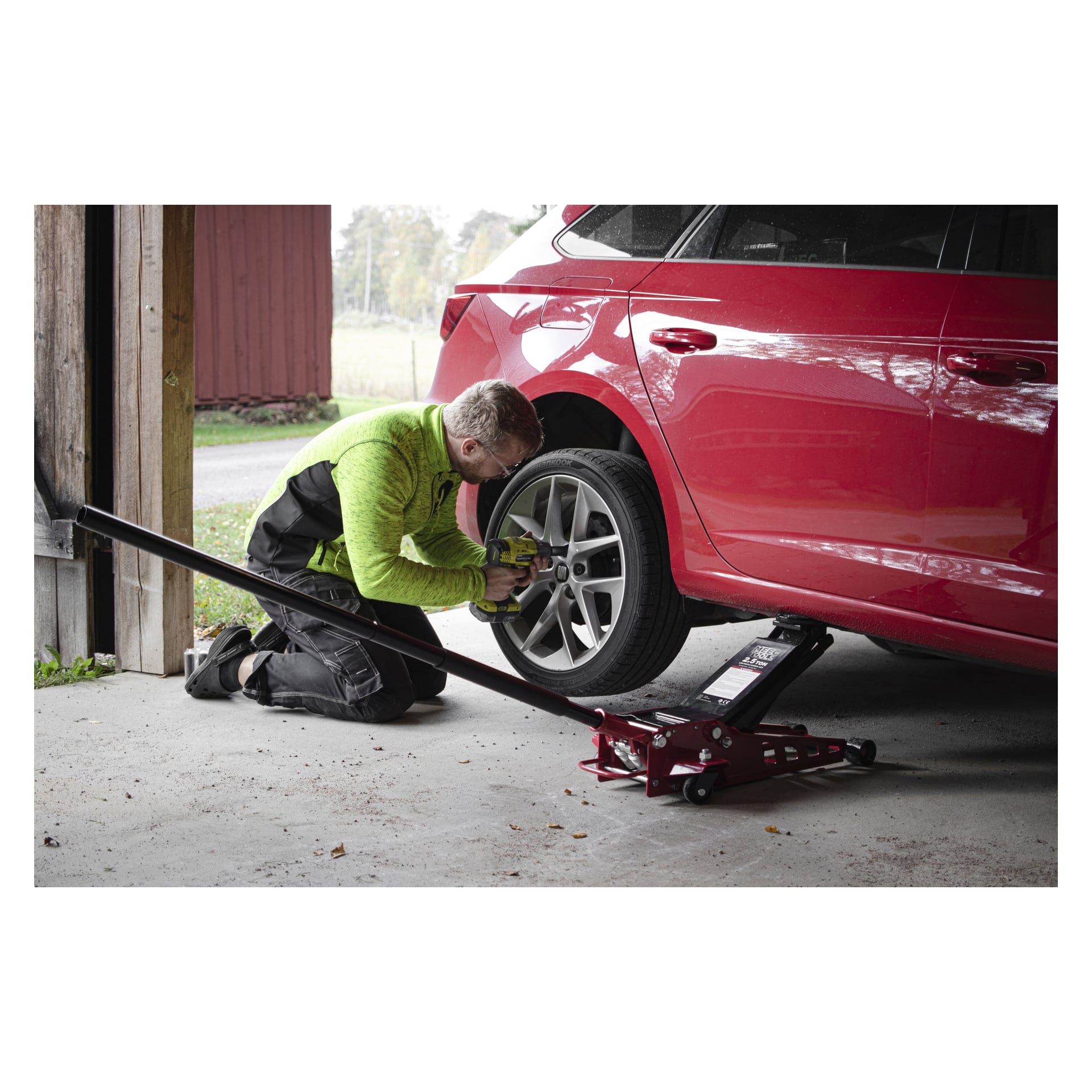 Eine Person in grüner Jacke und schwarzer Hose kniet auf dem Garagenboden und benutzt ein Werkzeug, um das Vorderrad eines roten Autos mit dem Meec Tools High Lifting Low Profile Jack zu lösen. Das offene Garagentor im Hintergrund zeigt eine Außenansicht.