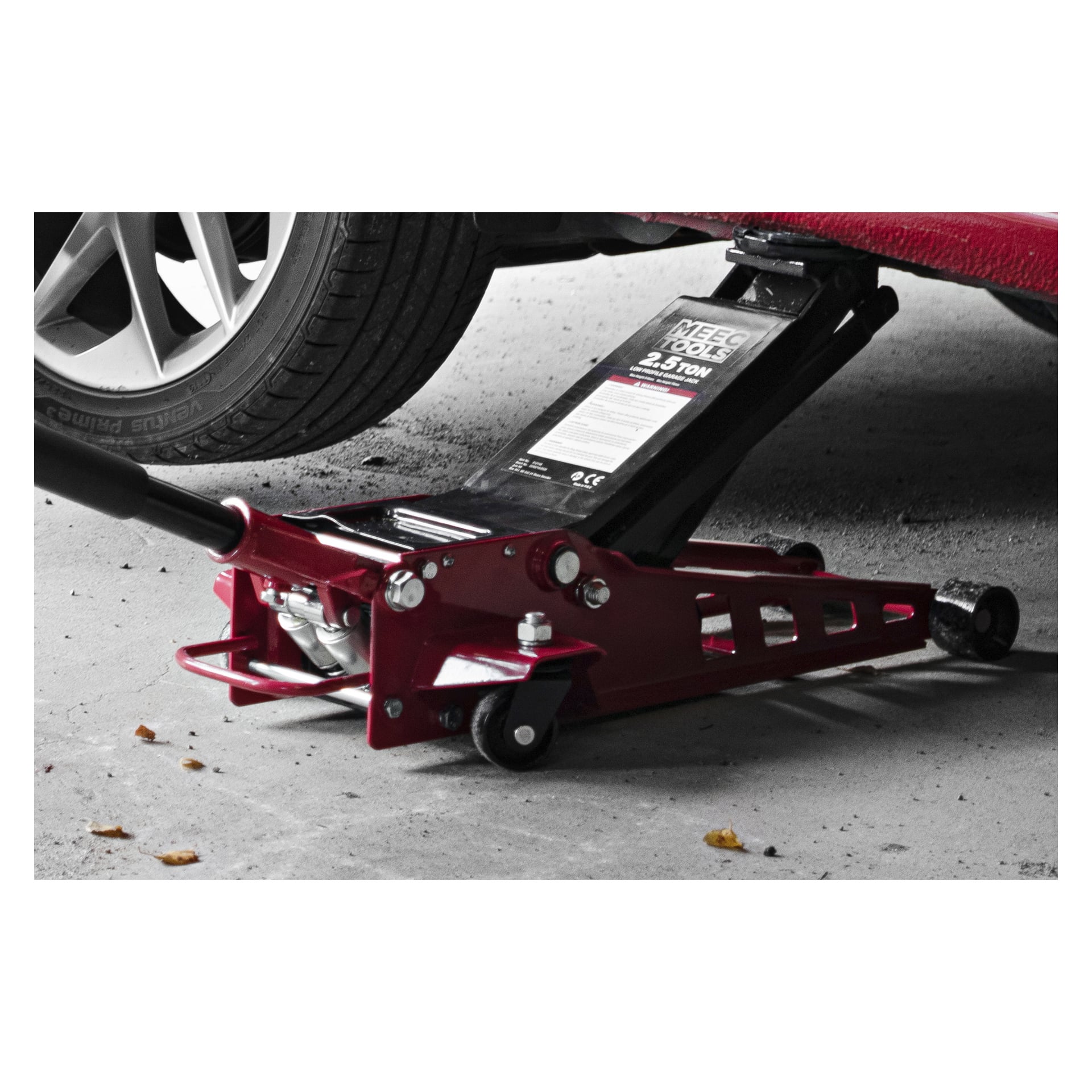 Ein roter Meec Tools High Lifting Low Profile Jack hebt ein Auto von der Seite an, wobei das Hinterrad und ein Teil des Fahrzeugs sichtbar sind. Das Etikett des Jacks zeigt an, dass er 2,5 Tonnen heben kann, und mehrere kleine Blätter liegen auf dem Boden daneben.