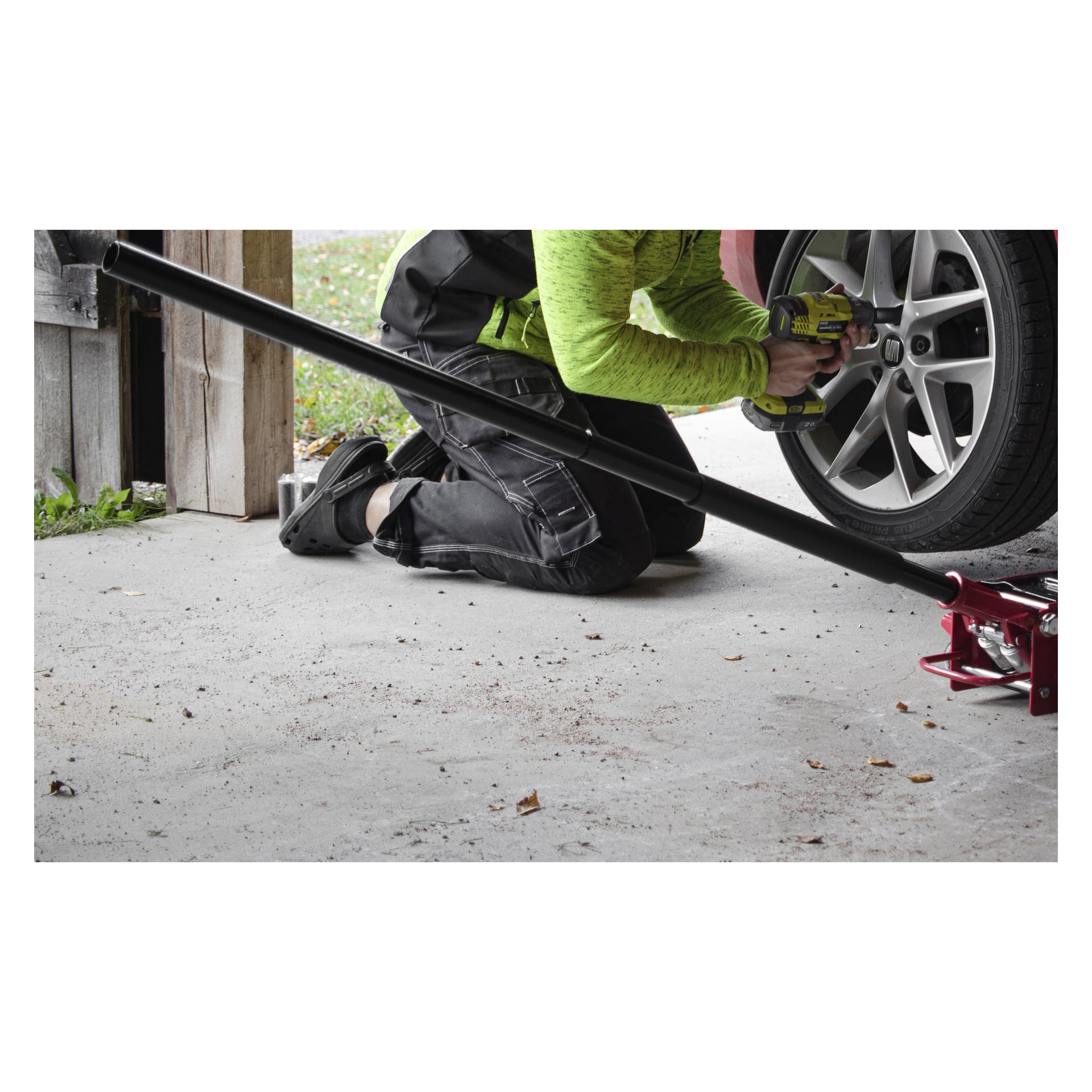 Mit einer grünen Jacke kniet eine Person auf dem Garagenboden und bedient ein Elektrowerkzeug am Vorderrad des Autos. Ein Meec Tools High Lifting Low Profile Jack sitzt unter dem Auto mit dem Griff zur Kamera gerichtet. Gefallene Blätter liegen auf dem Betonboden verstreut.