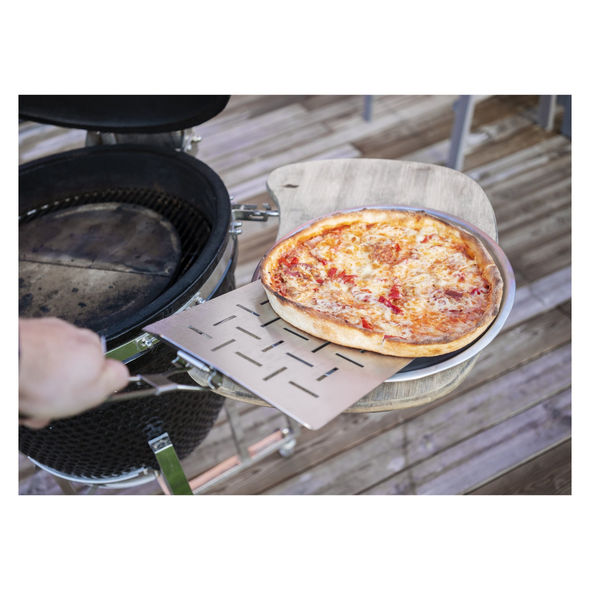 Ktoś podnosi świeżo upieczoną pizzę z serem i pomidorami z grilla za pomocą Burns & Barkles Pizza Spatula. Pizza stoi na okrągłej tacy, która jest ustawiona na drewnianej powierzchni obok grilla na tarasie.