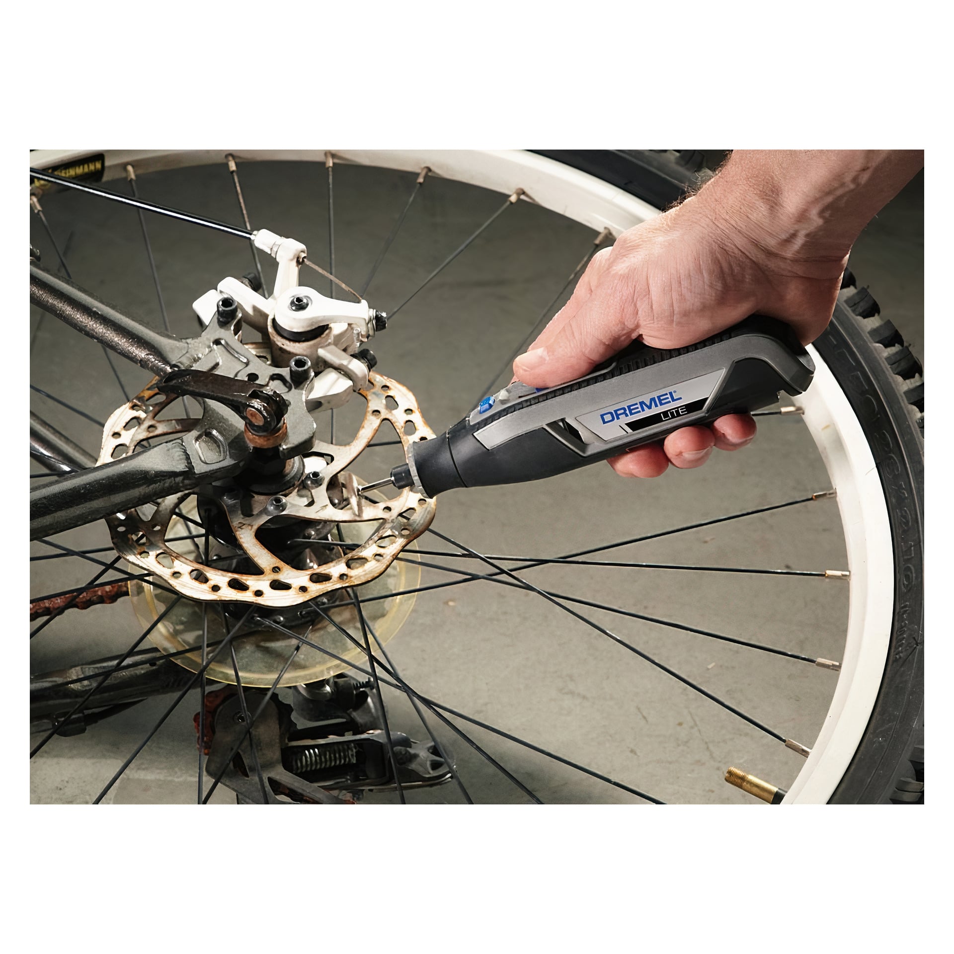 Någon använder ett Dremel Lite 7760-15 slip- och gravyrverktyg för att rengöra eller polera skivbromsskivan på en cykel. Enheten är placerad nära cykelns bakhjul, och rotorn ser rostig eller smutsig ut.