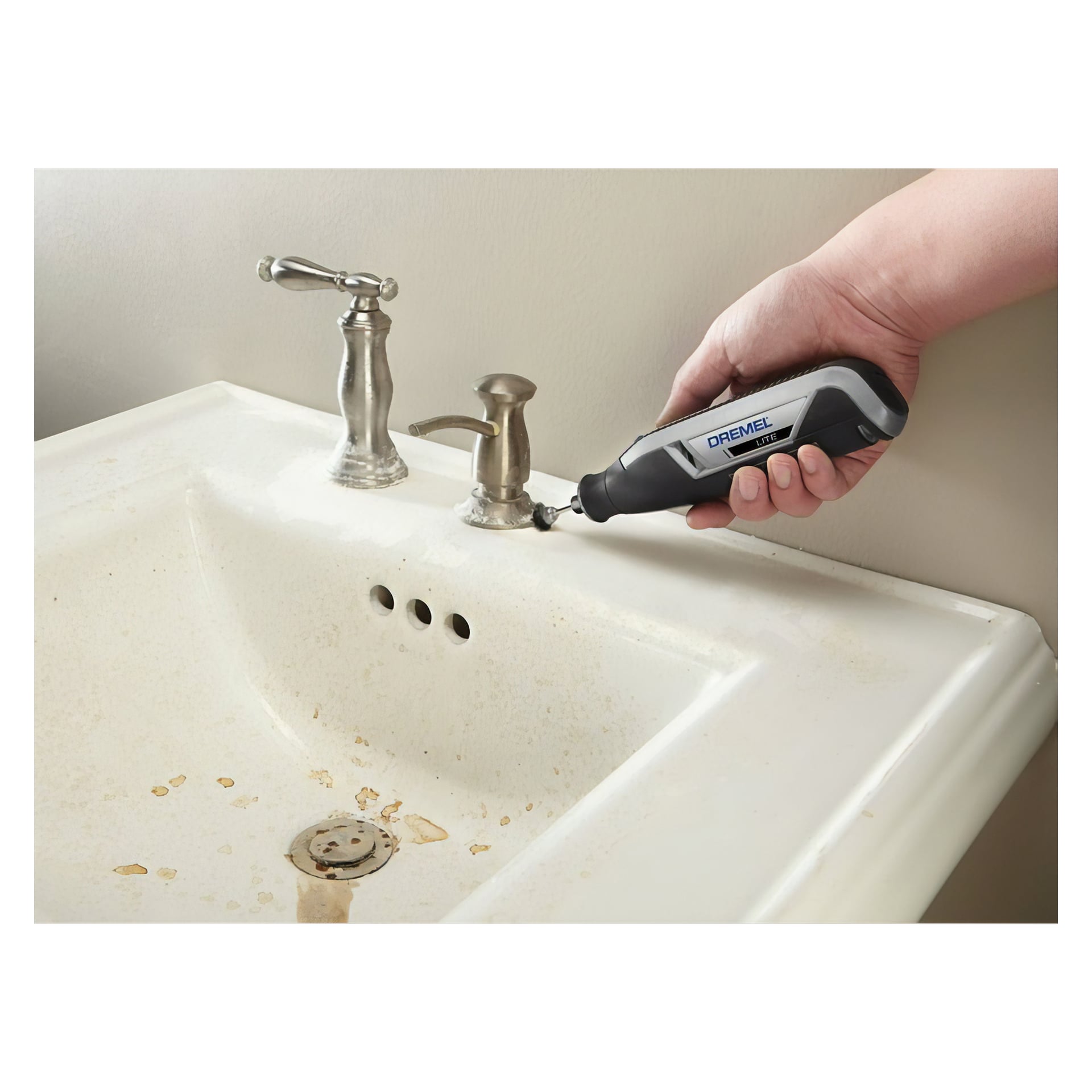Man använder ett svart och grått Dremel Lite 7760-15 slip- och gravyrverktyg för att ta bort smuts från basen på en kran på en fläckad vit badrumshandfat, med synlig rost.