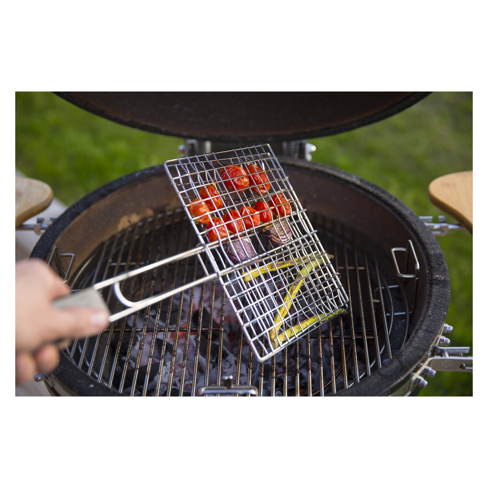 Man holder Burns & Barkles BBQ Grill-halsteren full av grønnsaker som tomater, rødløk og gul paprika, og holder den over en rund kullgrill med en metallrist utendørs på et gressområde.