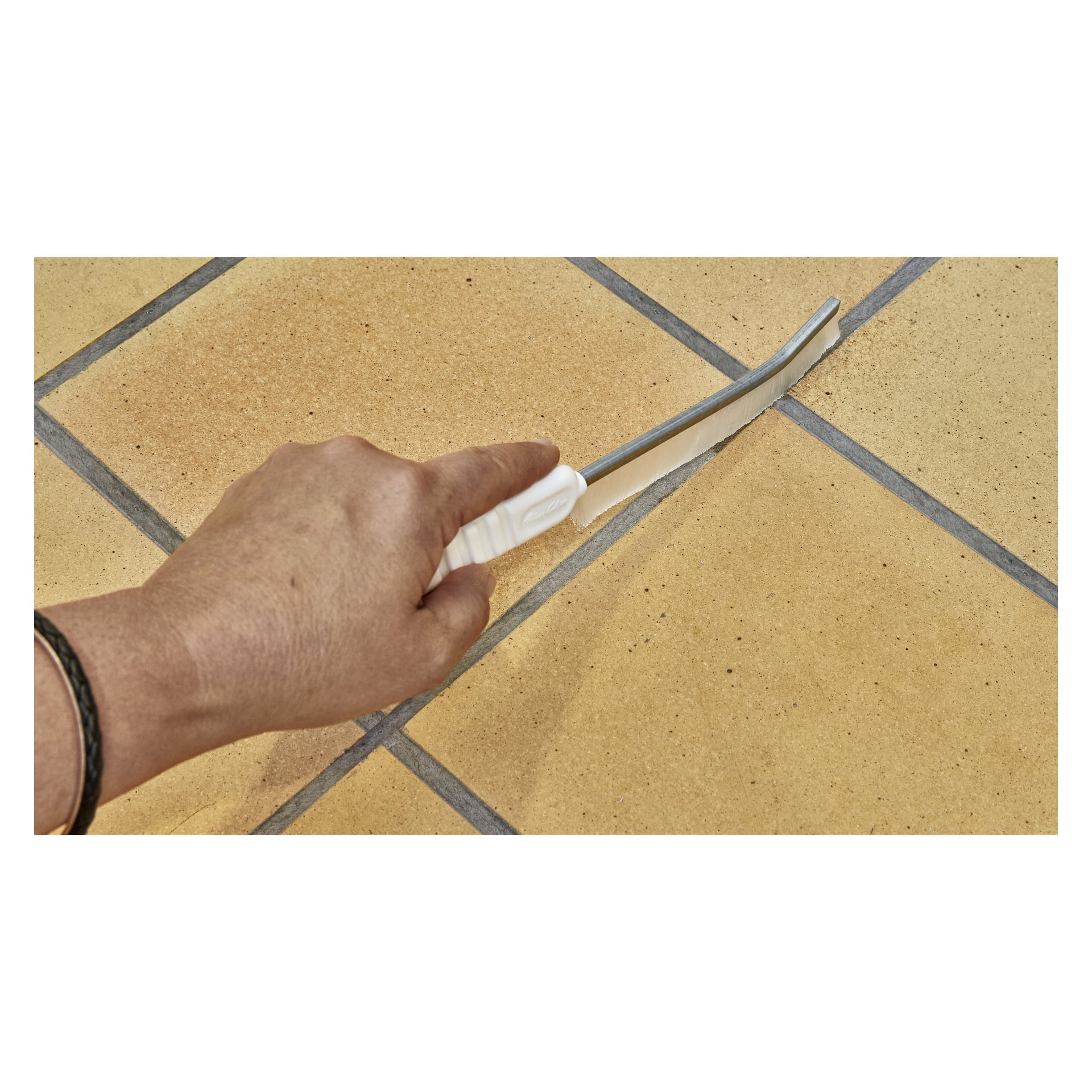 Ręka trzyma szczoteczkę Menuett Joint, czyszczącą fugę między jasnobrązowymi ceramicznymi płytkami z ciemnymi liniami fugi.