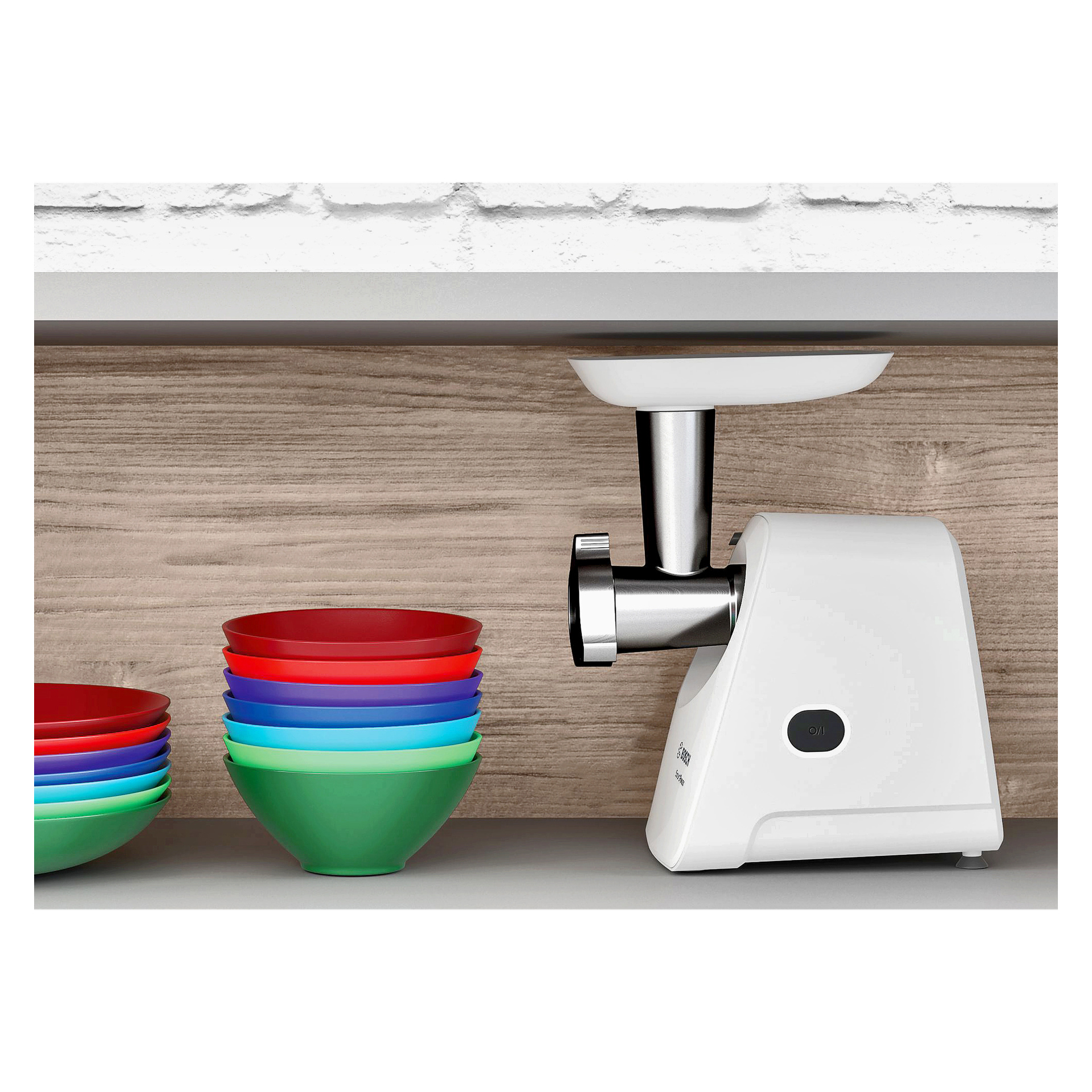 En Bosch Home Meat mincer MFW2510W i vitt står på en bänkskiva bredvid två staplar av färgstarka plastskålar i rött, blått och grönt, med en bakgrund av träådringar och vit tegelvägg ovanför.