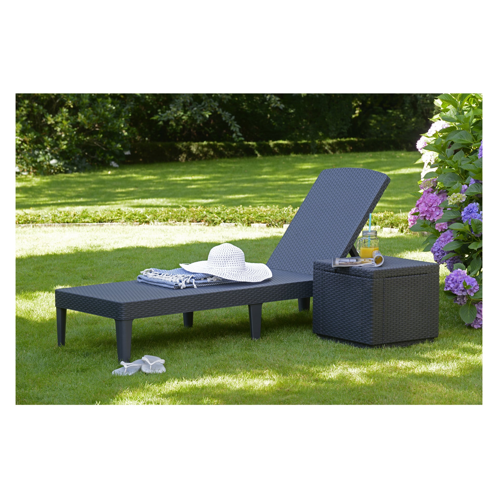 En solbädd i Keter Jaipur står på gräset bredvid ett matchande litet bord som rymmer en dryck, handduk, solhatt och sandaler. I bakgrunden finns träd och blommande lila blommor.