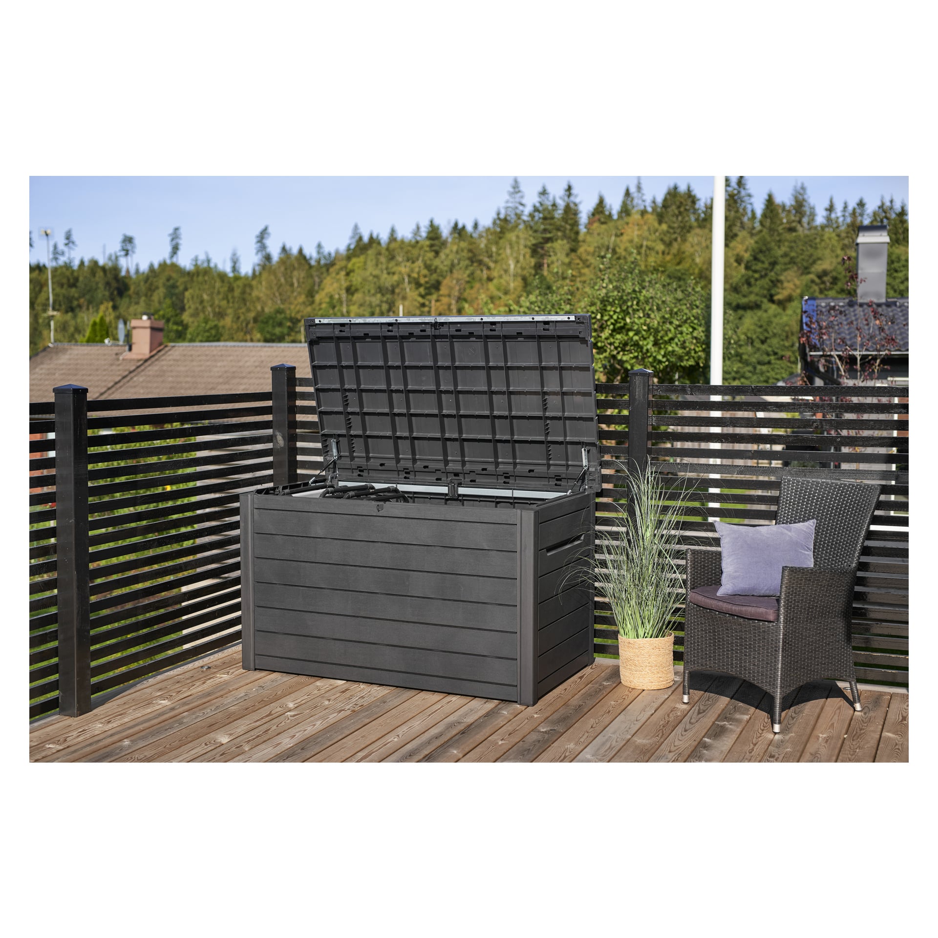 Keter Cushion Box Ontario vises med lokket åpent på en treterrasse. Ved siden av står en kurvstol med en pute og en potteplante. Bakgrunnen inkluderer trær og hus synlige bak rekkverket på terrassen.
