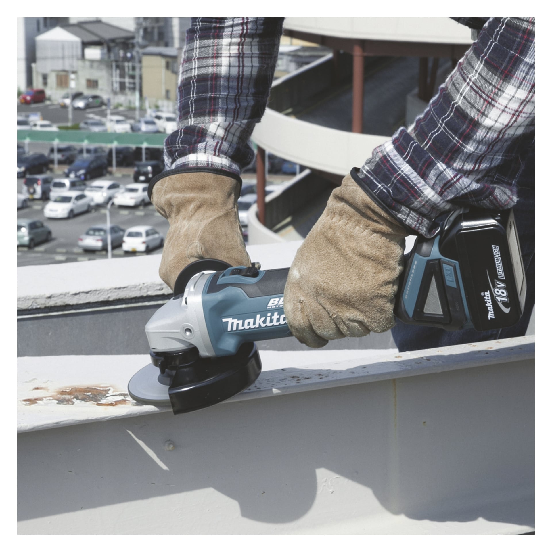 En person i rutiga ärmar och skyddshandskar använder en Makita vinkelslip DGA506Z och slipar rostigt metall utanför nära en parkeringsplats.