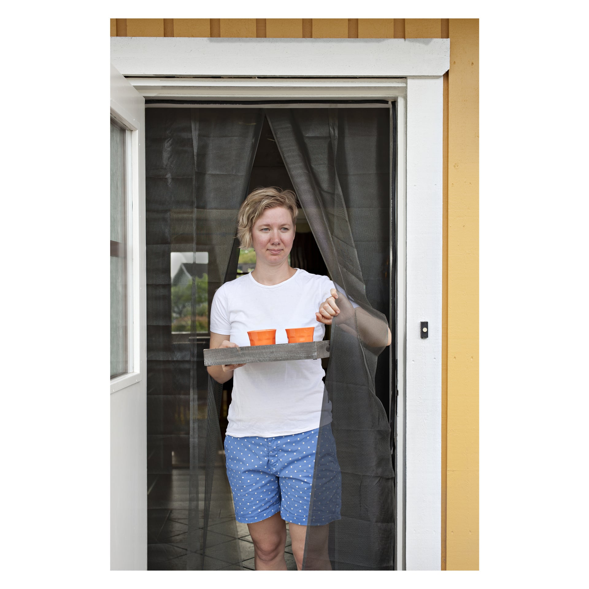 En person i vit skjorta och blå prickiga shorts står i en dörröppning och bär på en bricka med två orange koppar. Bakom dem hänger ett Hard Head-myggnät framför en gul vägg.