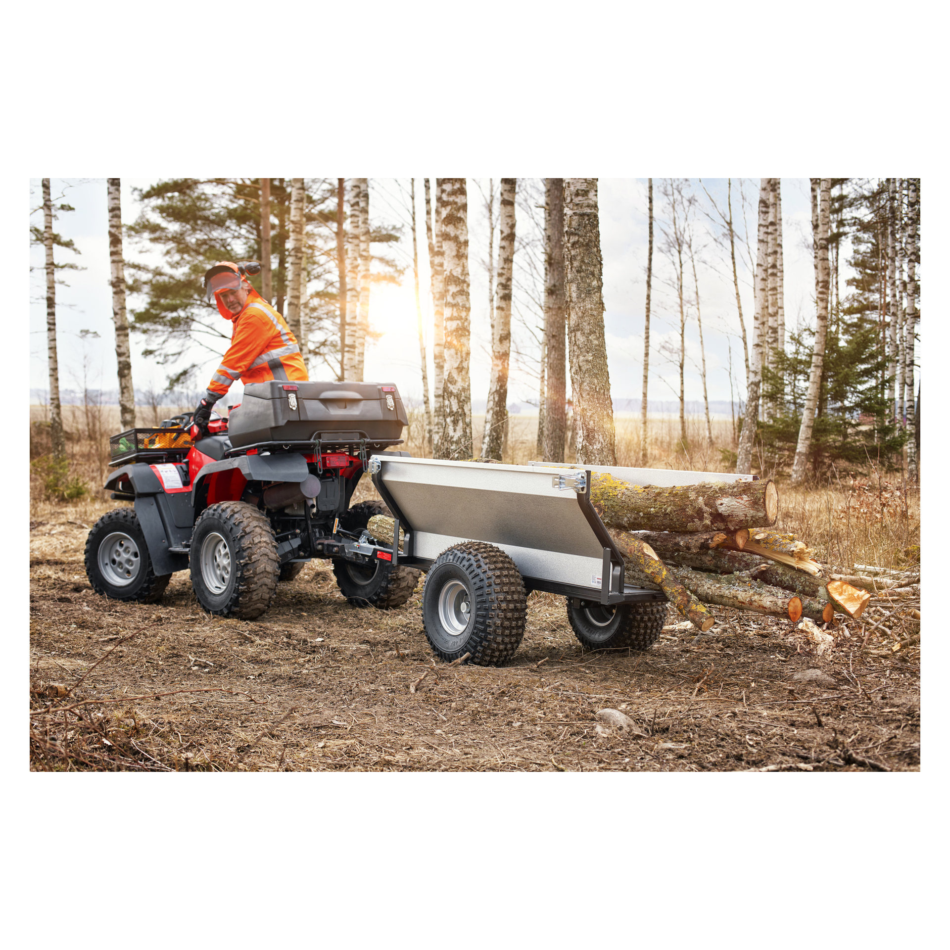 Med skyddsutrustning kör man en fyrhjuling med en Hamron-trailer fäst och drar huggna stockar nerför en jordstig i en skog medan solljuset silas genom träden.