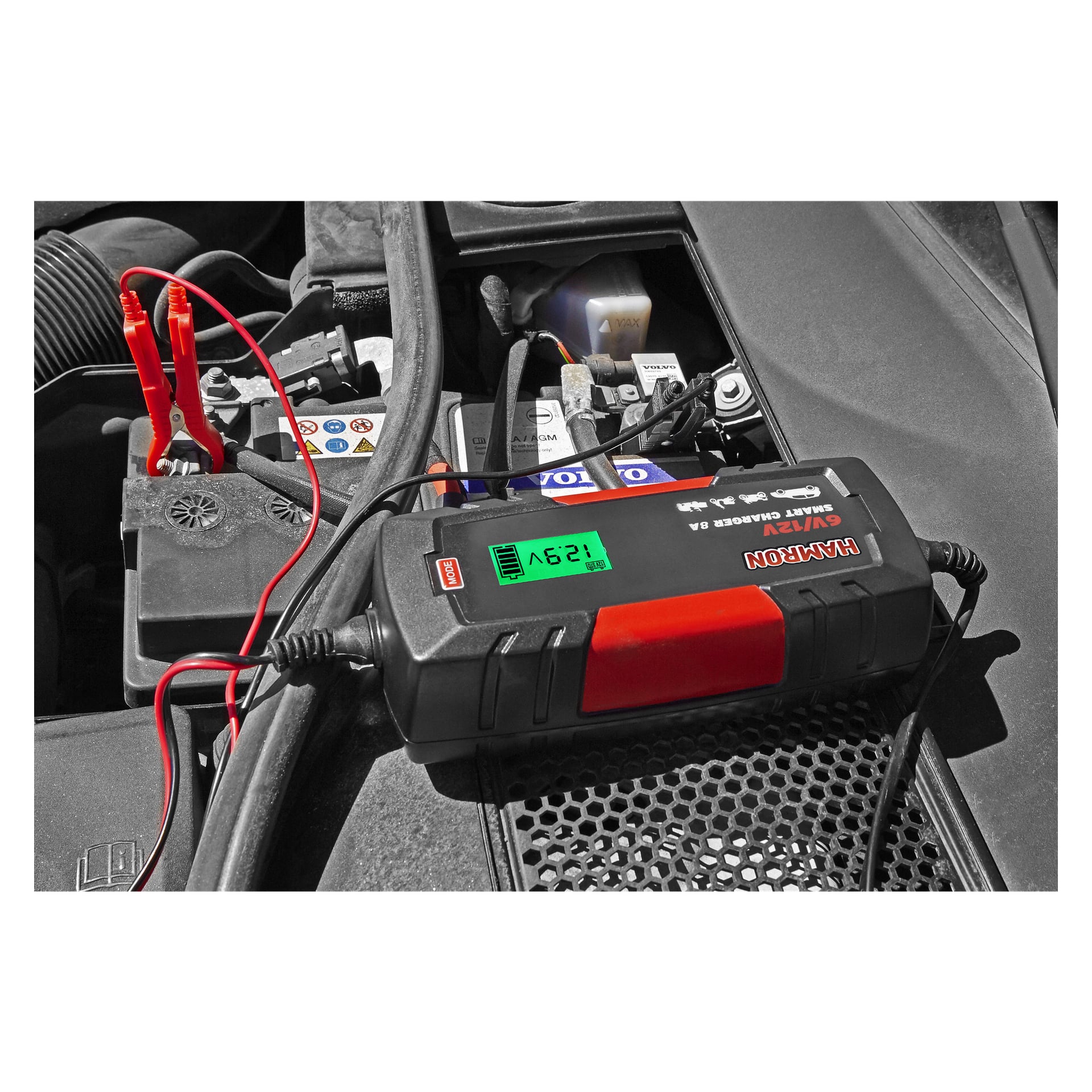 Et bilbatteri er koblet til en Hamron-batterilader med røde og svarte klemmer. Laderen viser 12,9V. Ulike batterideler og etiketter er synlige i bakgrunnen.