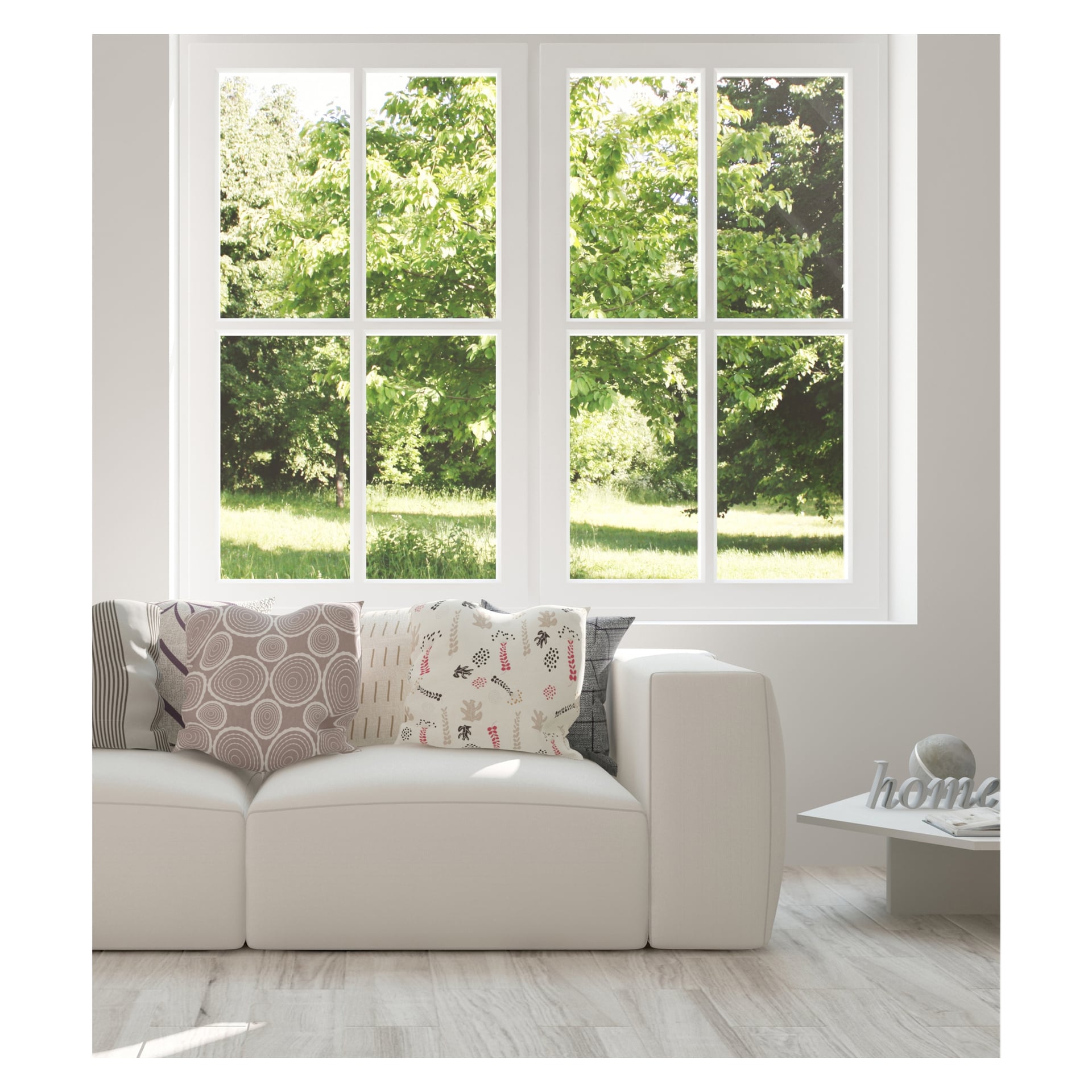 En moderne hvit sofa pyntet med mønstrede pynteputer er plassert foran store vinduer utstyrt med Hard Head vindusfilm, slik at sollyset lyser opp det frodige grøntområdet utenfor. Til høyre for sofaen står et lite sidebord med en dekorativ gjenstand som staver «home».