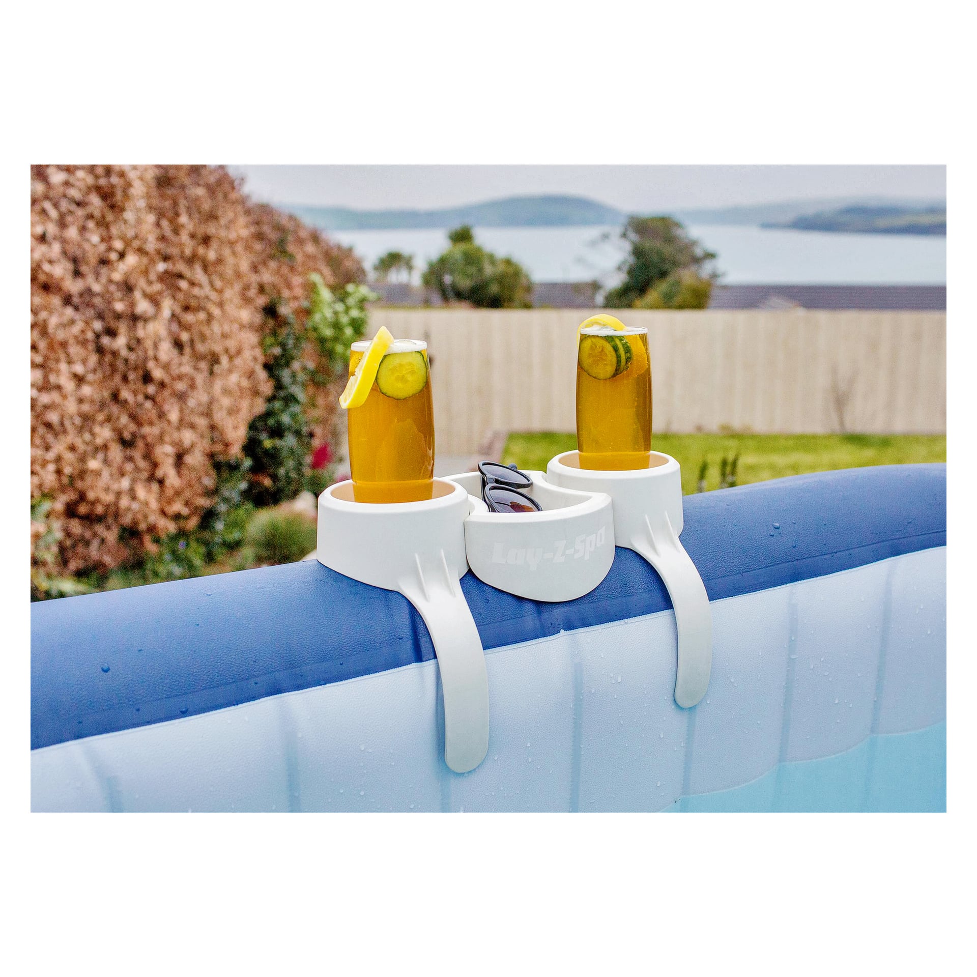 Der Bestway-Becherhalter für das Spa-Bad fasst zwei Getränke mit Zitronen- und Gurkenscheiben, die an einem aufblasbaren Whirlpool befestigt sind. Zwischen den Getränken sind Sonnenbrillen platziert, und im Hintergrund ist ein Garten mit Bäumen und Wasser zu sehen.