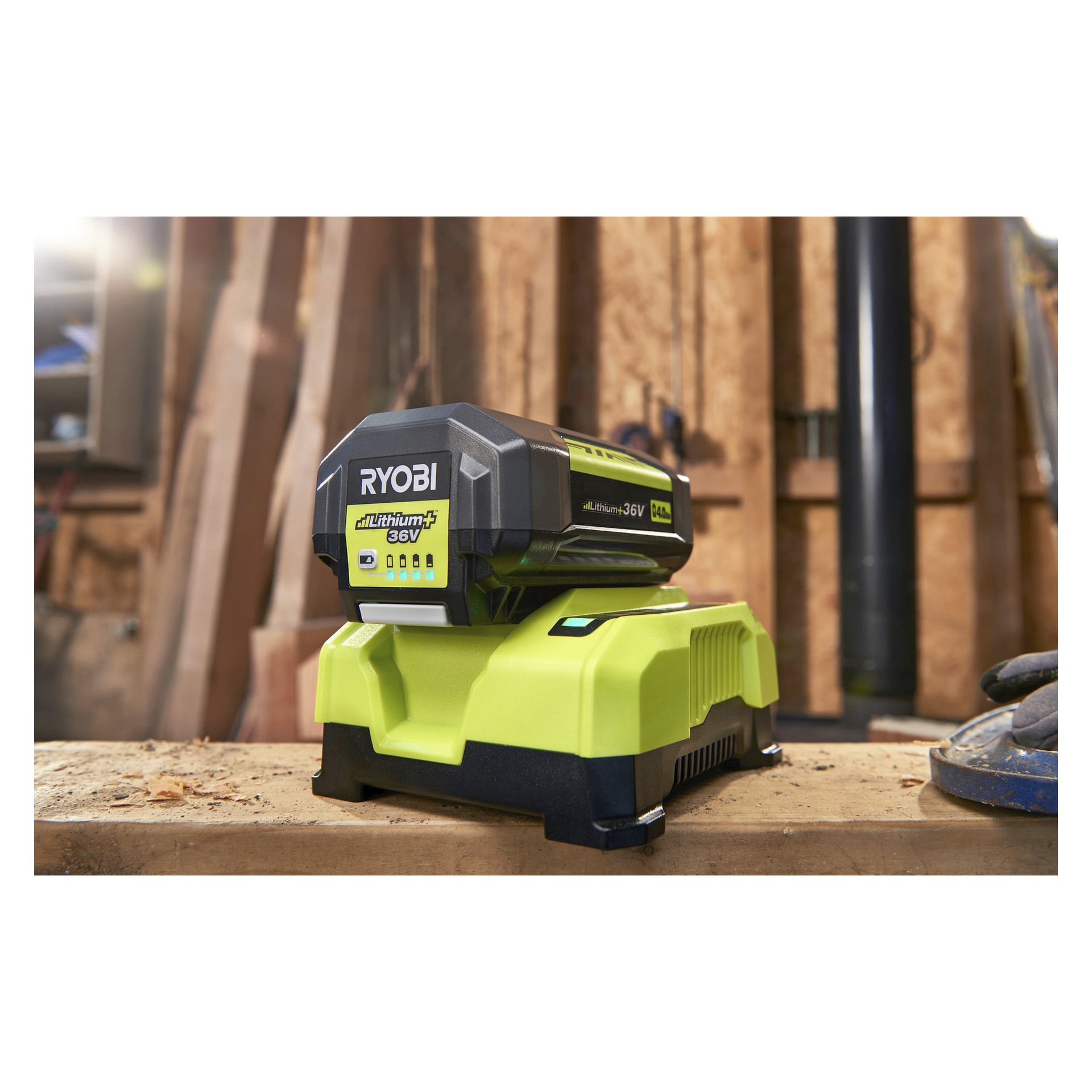 En Ryobi Max Power batterilader RY36C60A med et 36V litiumbatteri står plassert i et verksted omgitt av trebord og verktøy, og den livlige grønne fargen gjør den fremtredende på ladestasjonen.