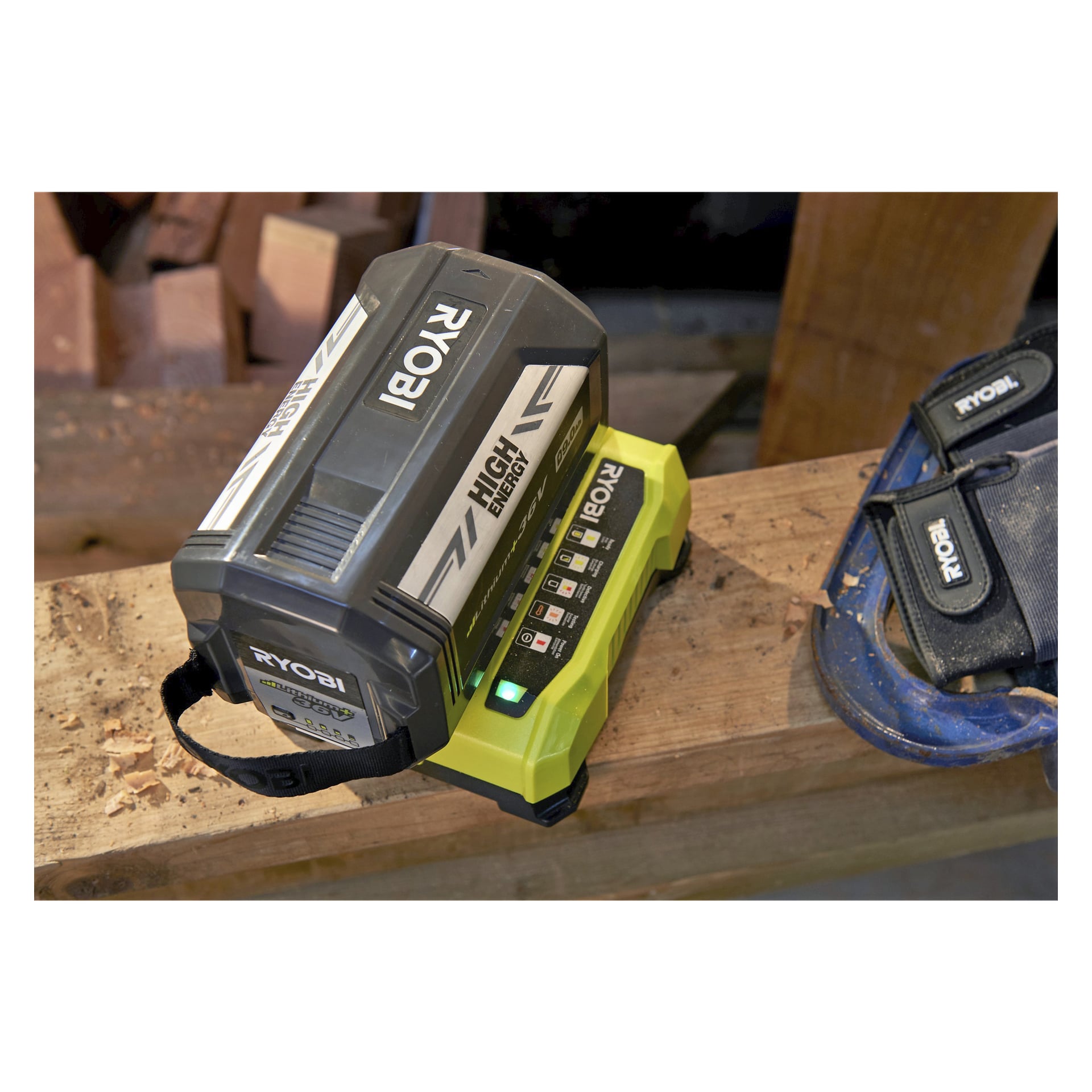 En Ryobi Max Power Battery-lader RY36C60A et høyenergibatteri på en trebjelke. I nærheten er det et blått verktøybelte og noen trebiter, og laderens indikatorlys lyser opp.