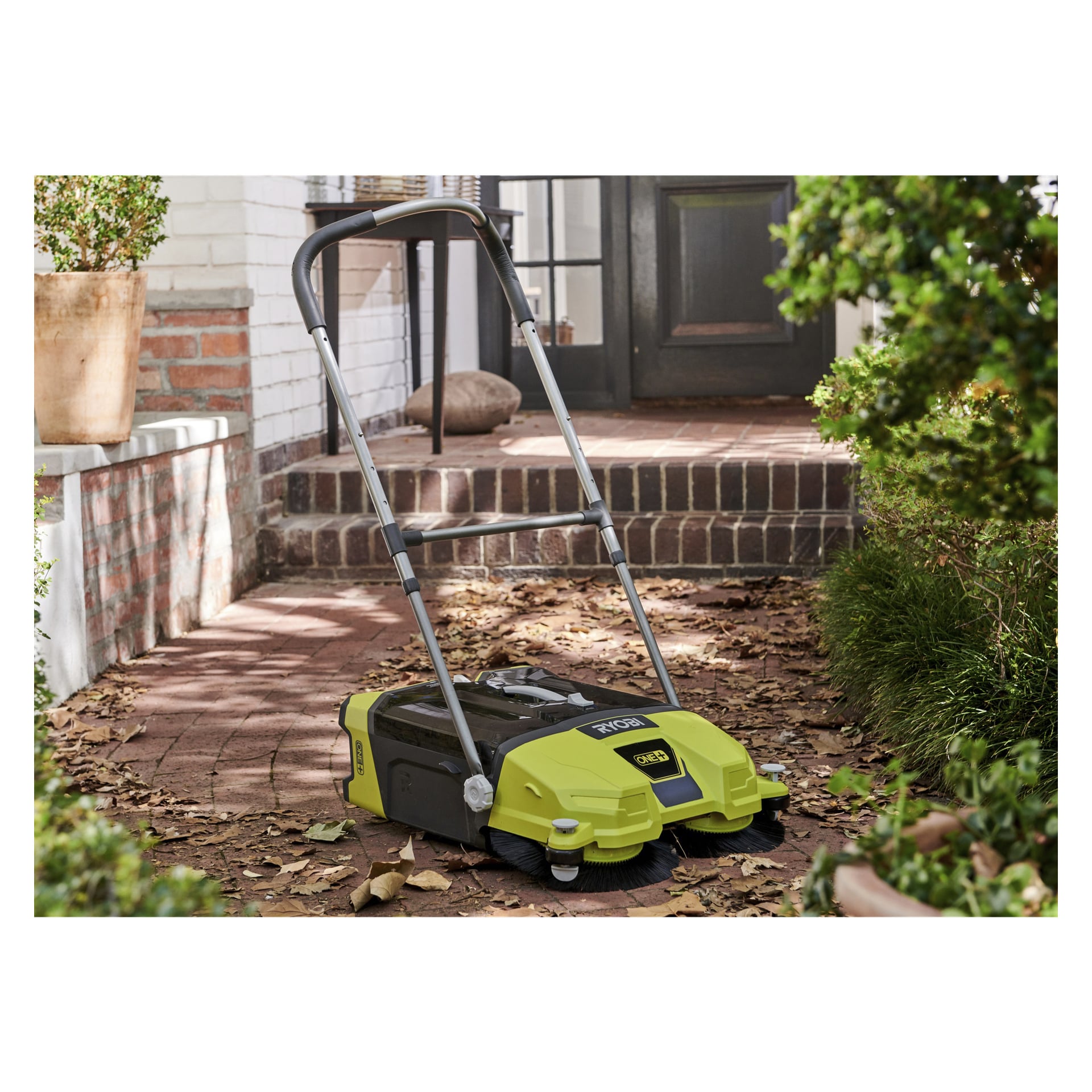 Der grün-schwarze Ryobi One+ Sweepers R18SW3-0 steht auf einem Backsteinweg mit trockenen Blättern, der an einer Haustür liegt, umgeben von Topfpflanzen und üppigem Grün.