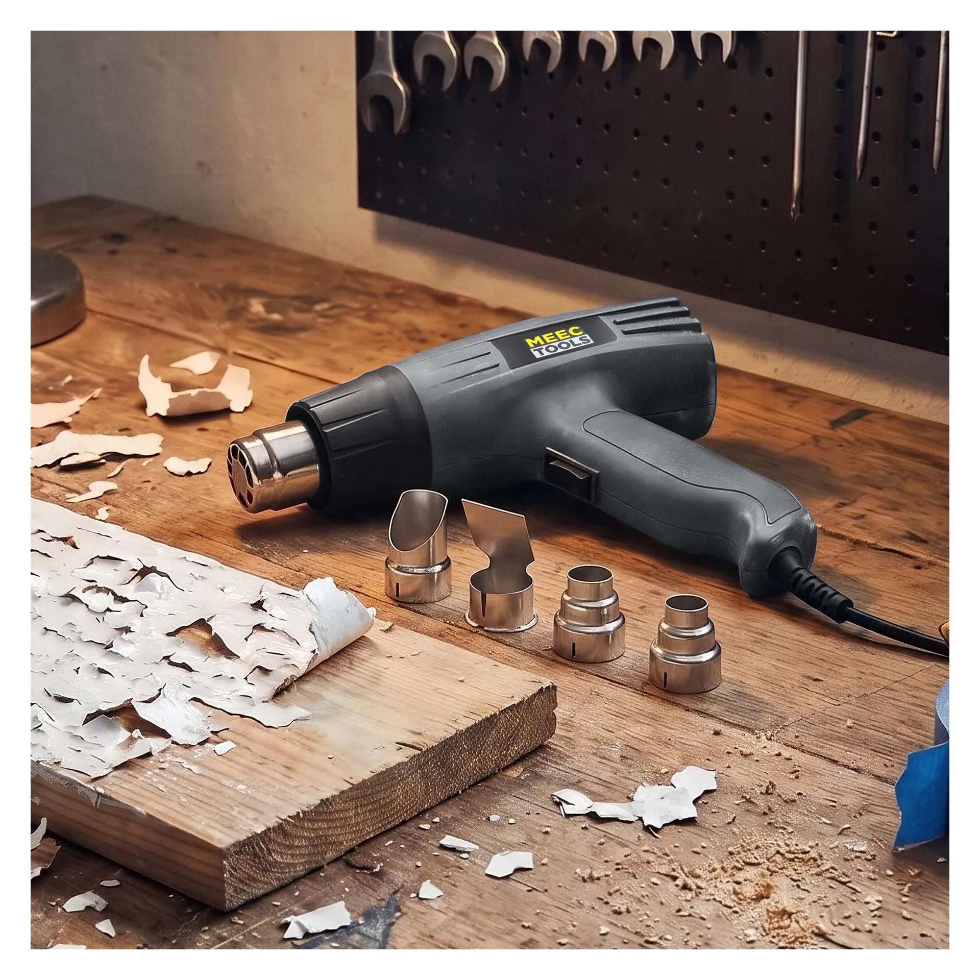 Musta Meec Tools Hot Air Gun on asetettu puiselle työpöydälle, joka on peitetty maalilaskeilla, neljän metallisuuttimen rinnalla. Taustalla naulataululla on useita käsityökaluja.