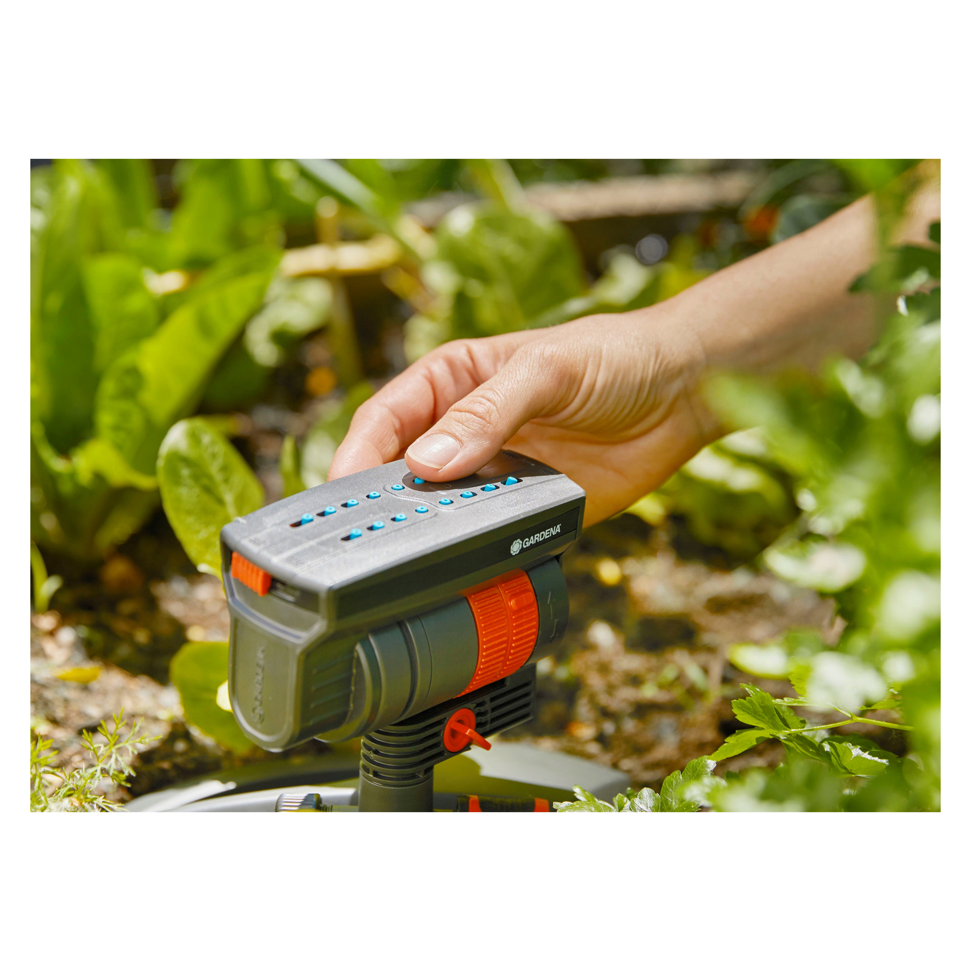 Käsi säätää Gardena AquaZoom Compact -kastelujärjestelmää puutarhassa, jonka takana on vihreitä kasveja.