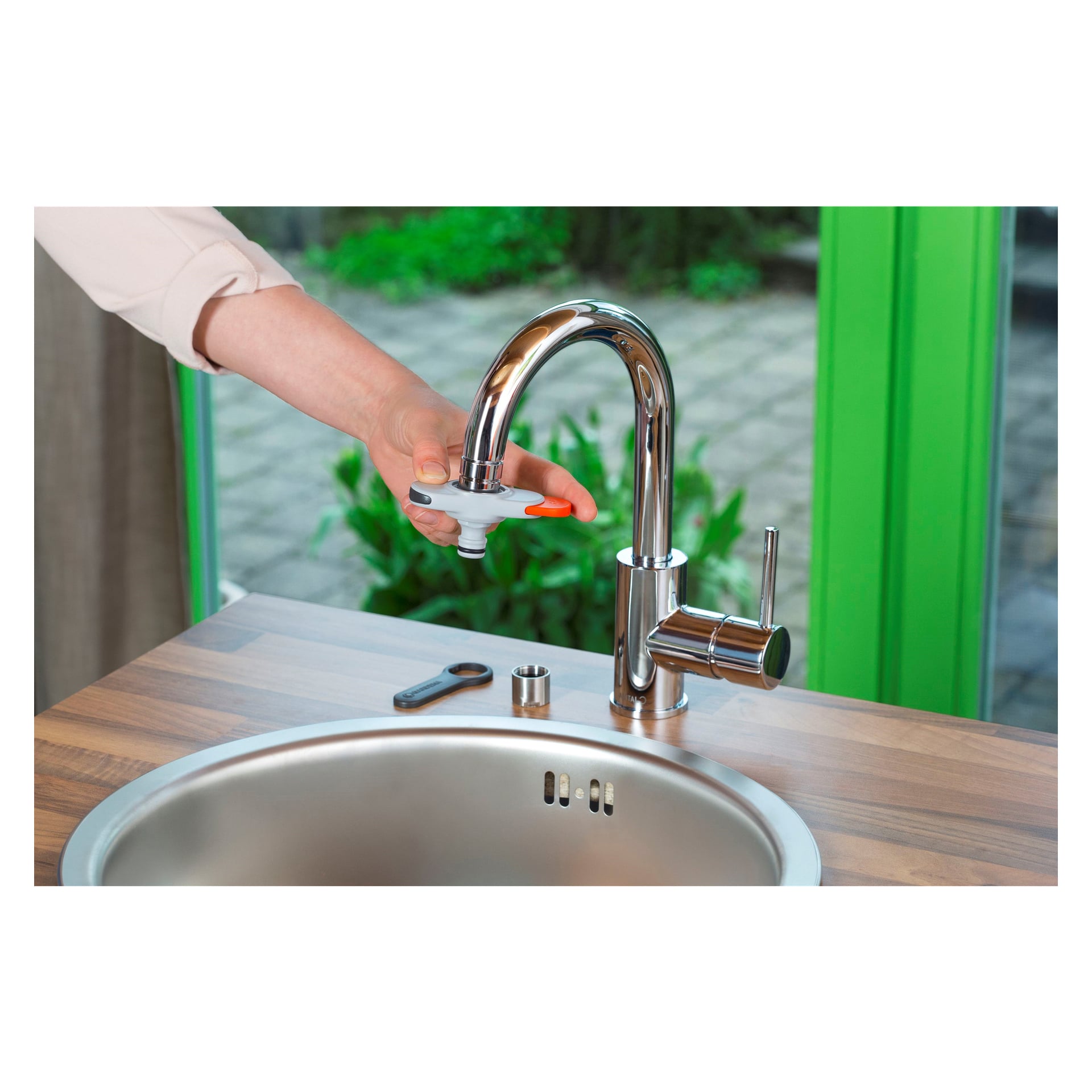 Noen fester eller kobler fra en Gardena Mixer-adapter på en skinnende krom kjøkkenkran. Vasken i rustfritt stål er synlig, og en skiftenøkkel samt flere krankomponenter ligger på trebenken. I bakgrunnen avslører grønne vinduer en utendørs terrasse.