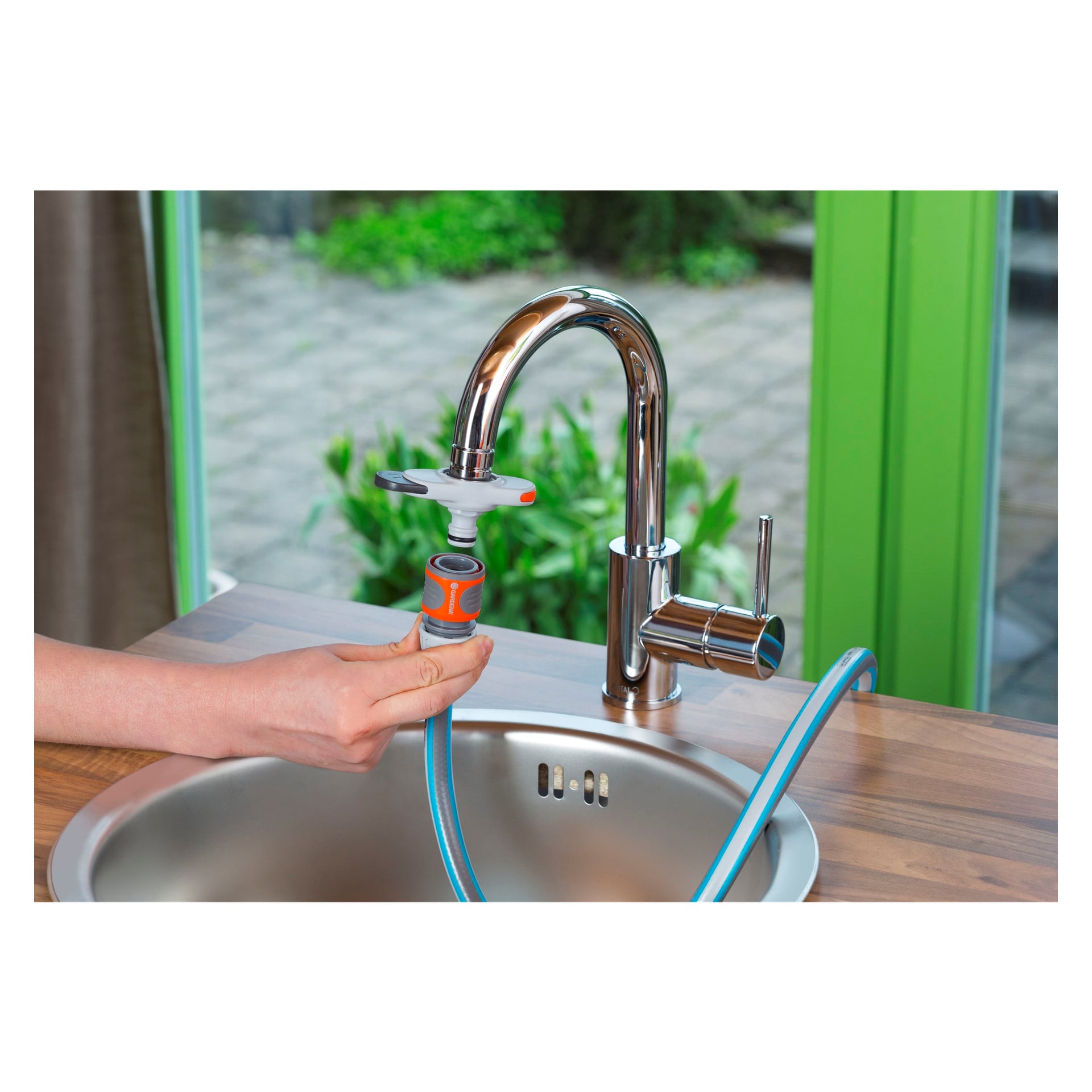 Noen installerer en Gardena Mixer-adapter på en kjøkkenkran i et stilrent, moderne kjøkken med vask i rustfritt stål. Utenfor vinduet kan du se en terrasse og frodig vegetasjon.