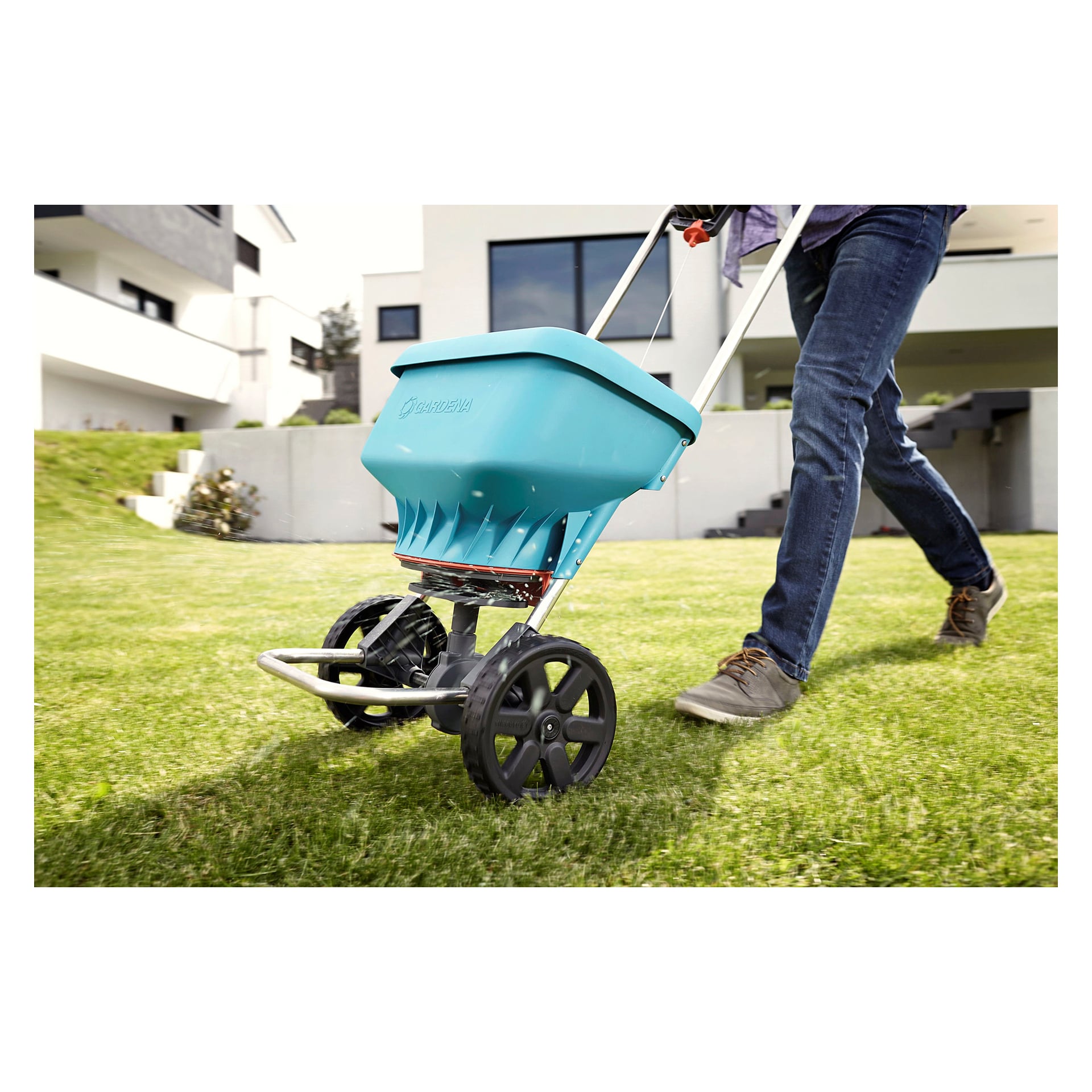 En person flytter en blå Gardena Universal-spreder over plenen i en forstadshage, med moderne hus og utetrapper synlige bak seg.