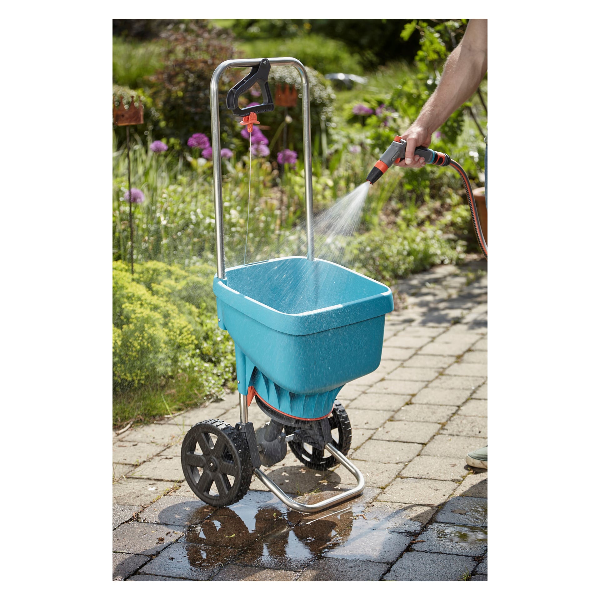 Noen skyller ned en blå Gardena Universal-spreder med svarte hjul på en steinterrasse, omgitt av grønne planter og blomstrende blomster i bakgrunnen.