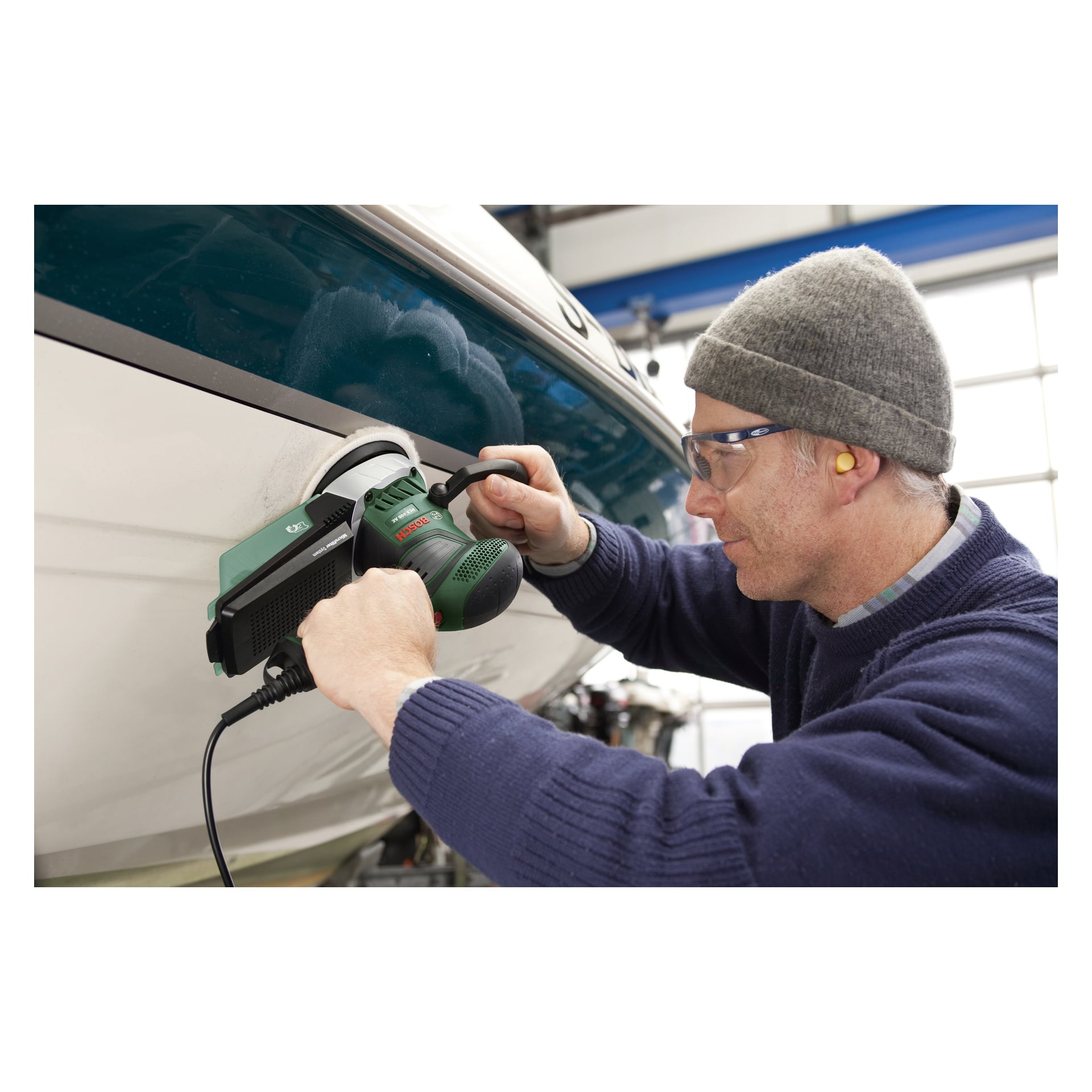 En person med skyddsglasögon, mössa och öronproppar slipar en båtsida med en Bosch Eccentric slipmaskin PEX 400 AE i en verkstad.