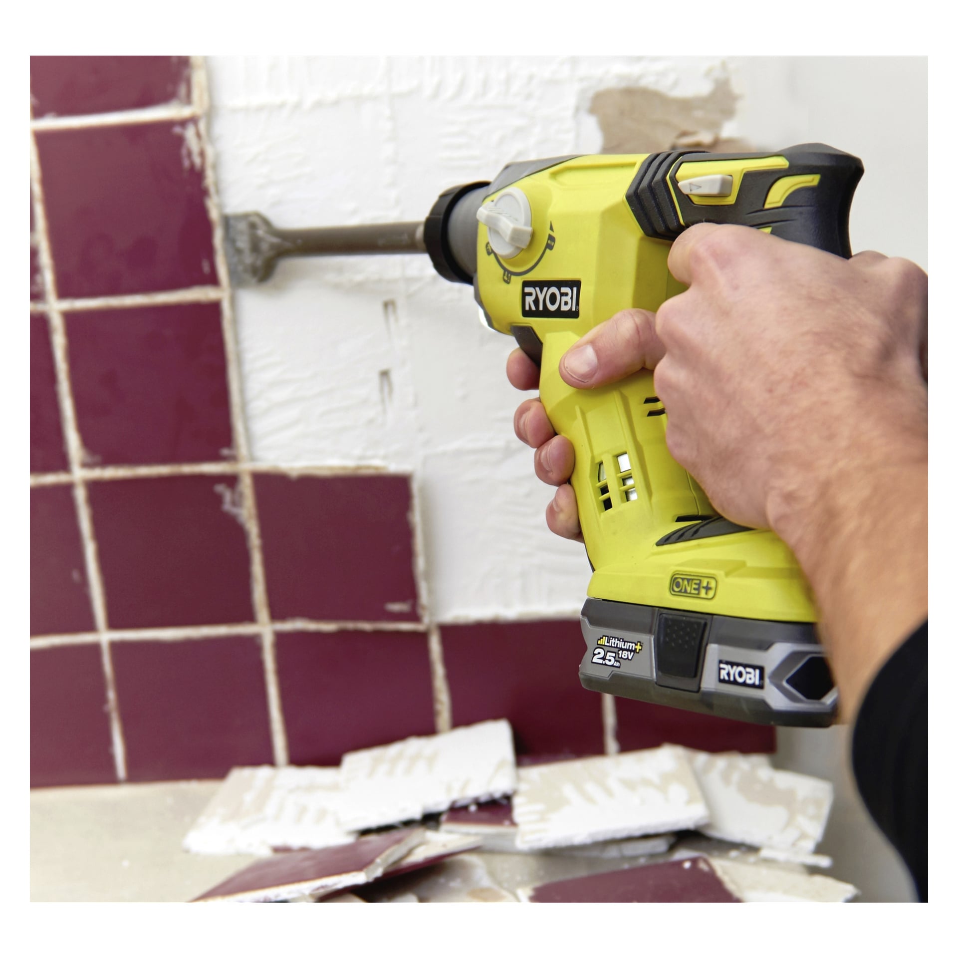 Man använder en Ryobi One+ R18SDS-0 batteridriven hammarborr med mejsel för att bända loss vinröda väggplattor. Bitar av trasiga plattor och fläckar av lim ligger utspridda på den vita väggen och bänkskivan under.