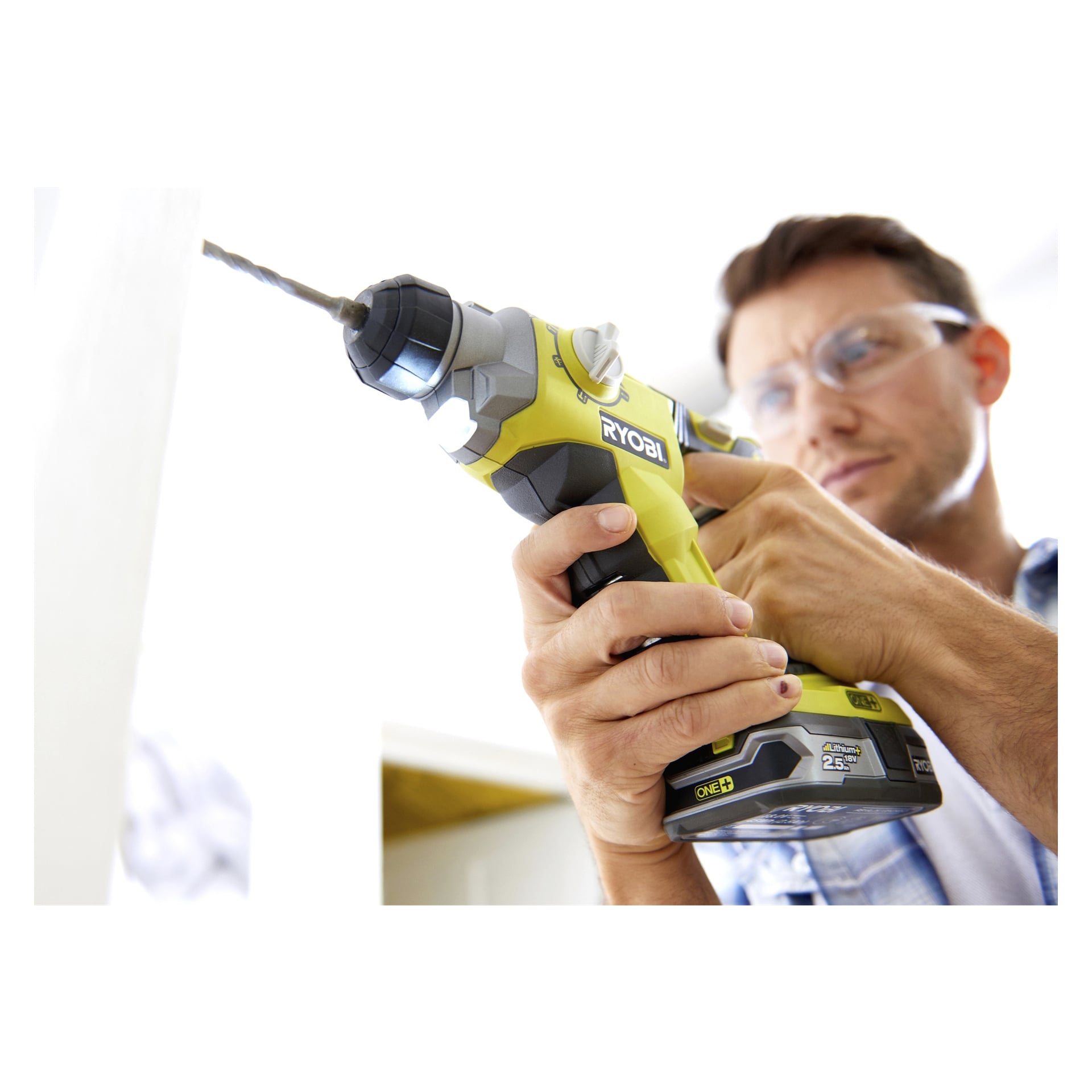 En person med skyddsglasögon fokuserar på att använda en Ryobi One+ R18SDS-0 batteridriven hammarborr med borr, och arbetar på en ljus inomhusyta.