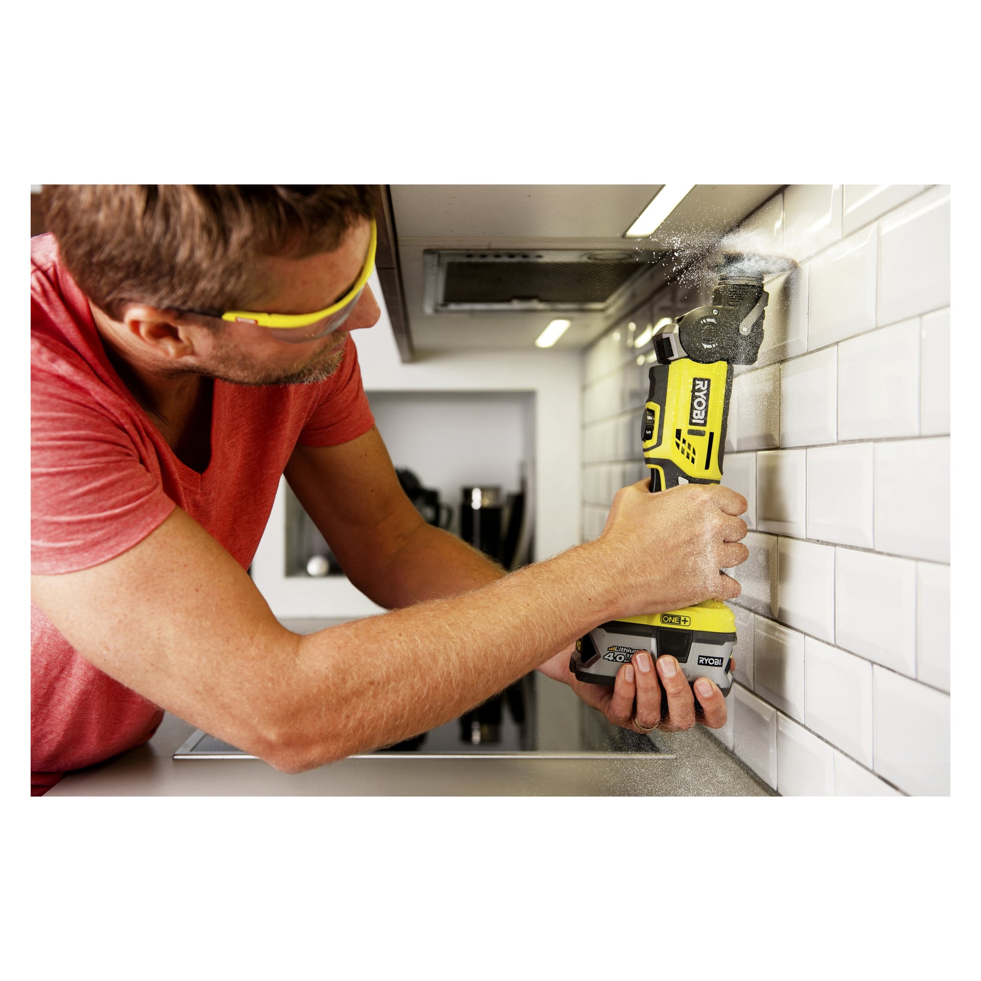 En person med skyddsglasögon använder en gul och svart Ryobi One+ Multitool R18MT-0 för att skära vita tunnelbaneplattor på en köksvägg, med damm synligt från skärningen.