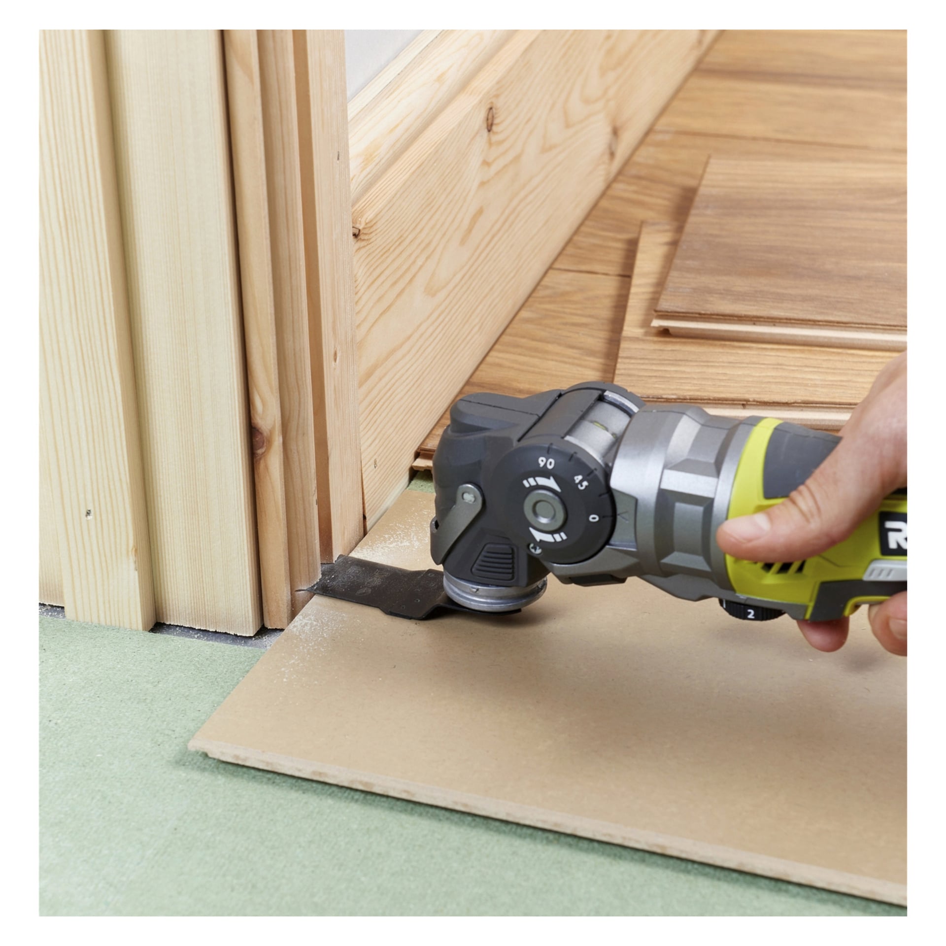 Någon använder en Ryobi One+ Multitool R18MT-0 för att trimma den nedre delen av en trädörrkarm, med en bit kartong placerad under för att skydda golvet.