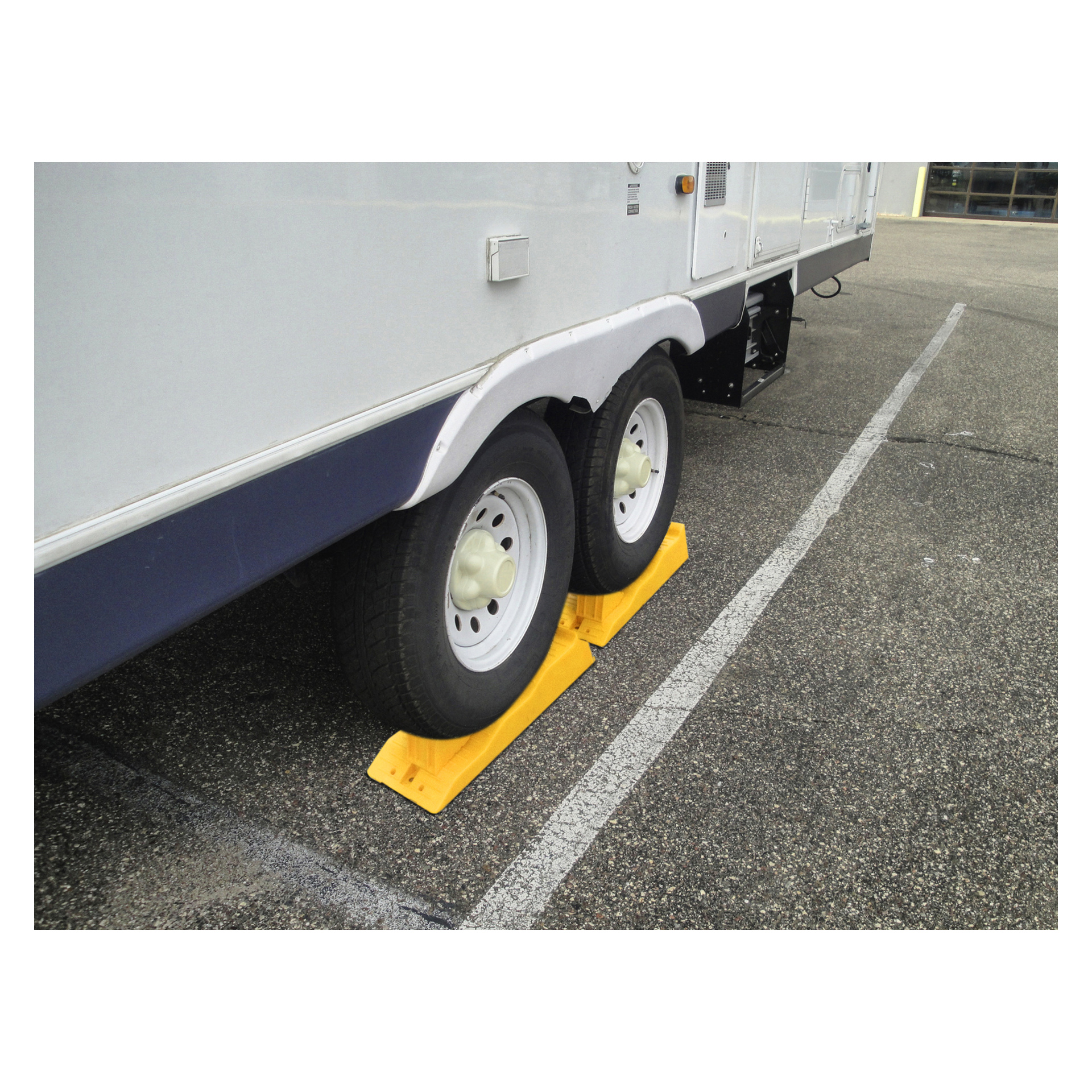 En vit trailer är parkerad på en parkering, med Hamron Level Blocks 2 Pack placerade på båda sidor av varje hjul för stabilitet. Trailern är placerad på parkeringsplatser markerade med vita linjer.