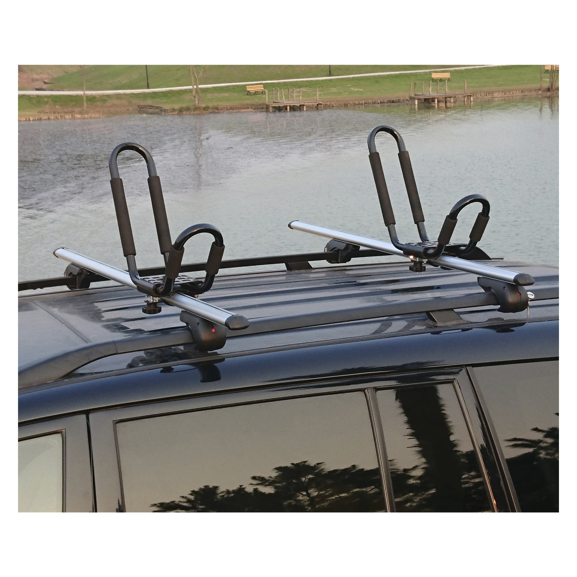 En närbild visar två Hamron Kayak Carrier 2 Pack-enheter monterade på taket av ett svart fordon, som står parkerat vid en vattenmassa med gräskanter och träbänkar i bakgrunden.