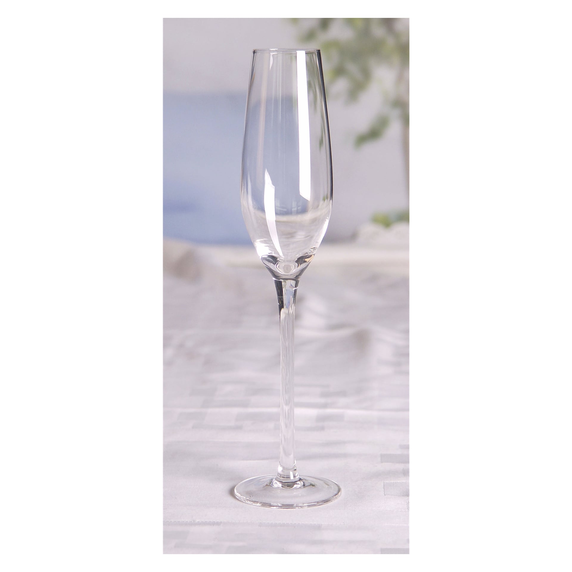 Ett klart champagneglas med lång stjälk från Menuett-setet med två är placerat på en lätt mönstrad bordsduk. Bakgrunden är mjukt ur fokus och visar inslag av grön växtlighet och blå toner som framkallar en lugn inomhusatmosfär.