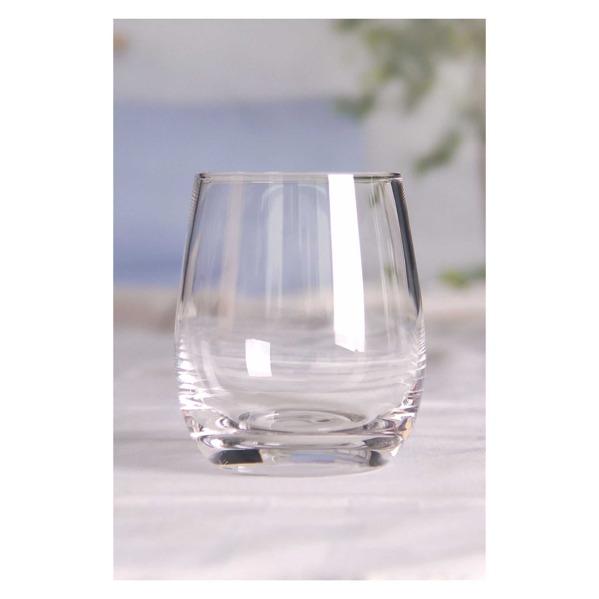 Ett klart dryckesglas från Menuett 2-paketet står på en yta, med en mjukt suddig bakgrund med grön växtlighet och blå toner som framkallar en lugn och ren atmosfär.