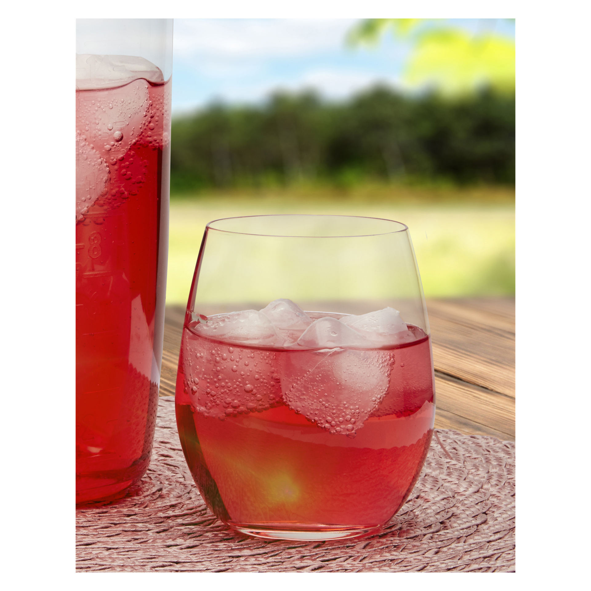 Ett Menuett-glas från tvåpacket placeras bredvid en kanna med en röd iced drink med isbitar på en rund vävd bordsunderlägg, med en suddig bakgrund av träd och gräs utomhus.