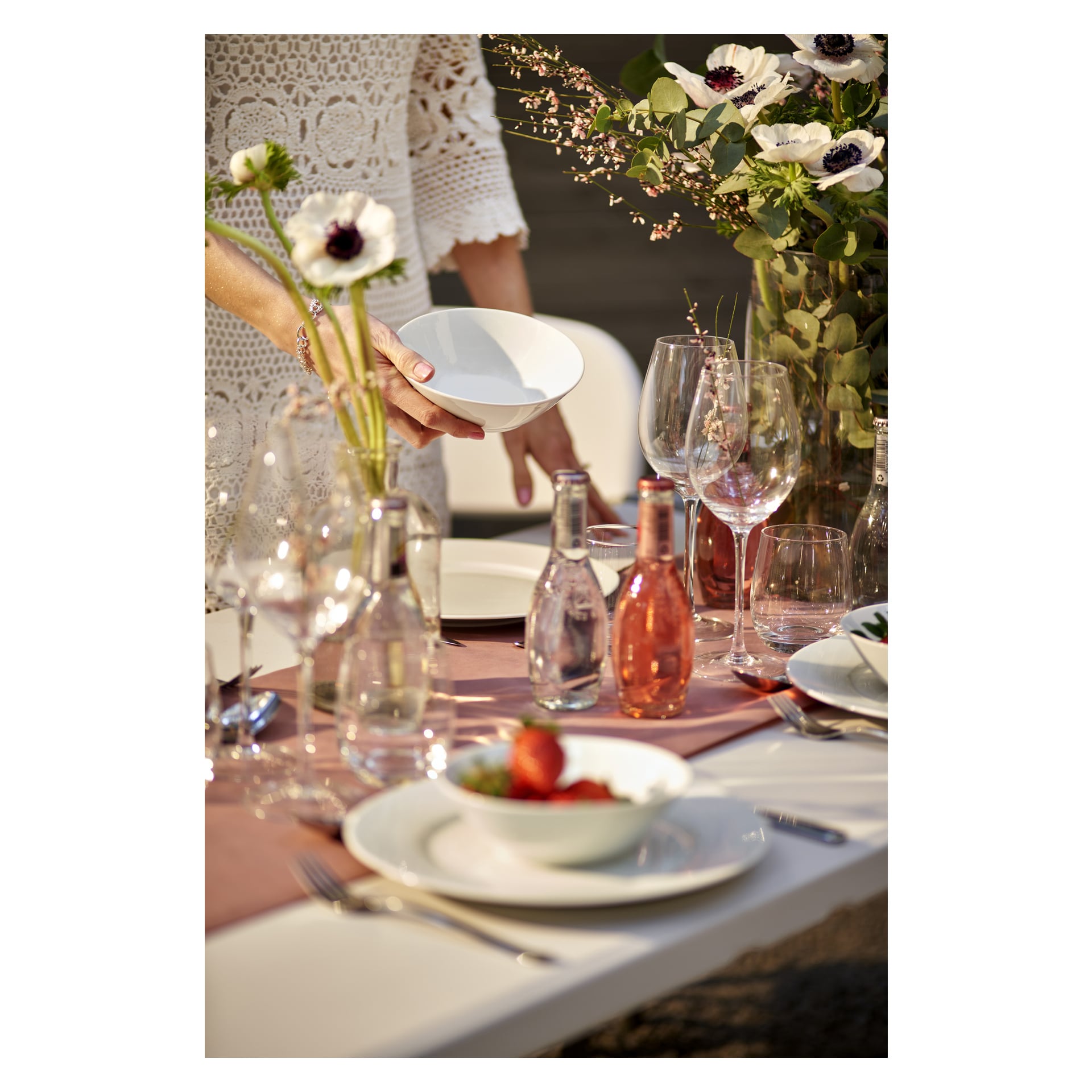 En person i en vit spetsklänning ställer en vit tallrik på ett utomhusbord prytt med rosa blommor, Menuett Drinking glass 2-pack, flaskor och skålar med mat, allt upplyst av naturligt solljus.