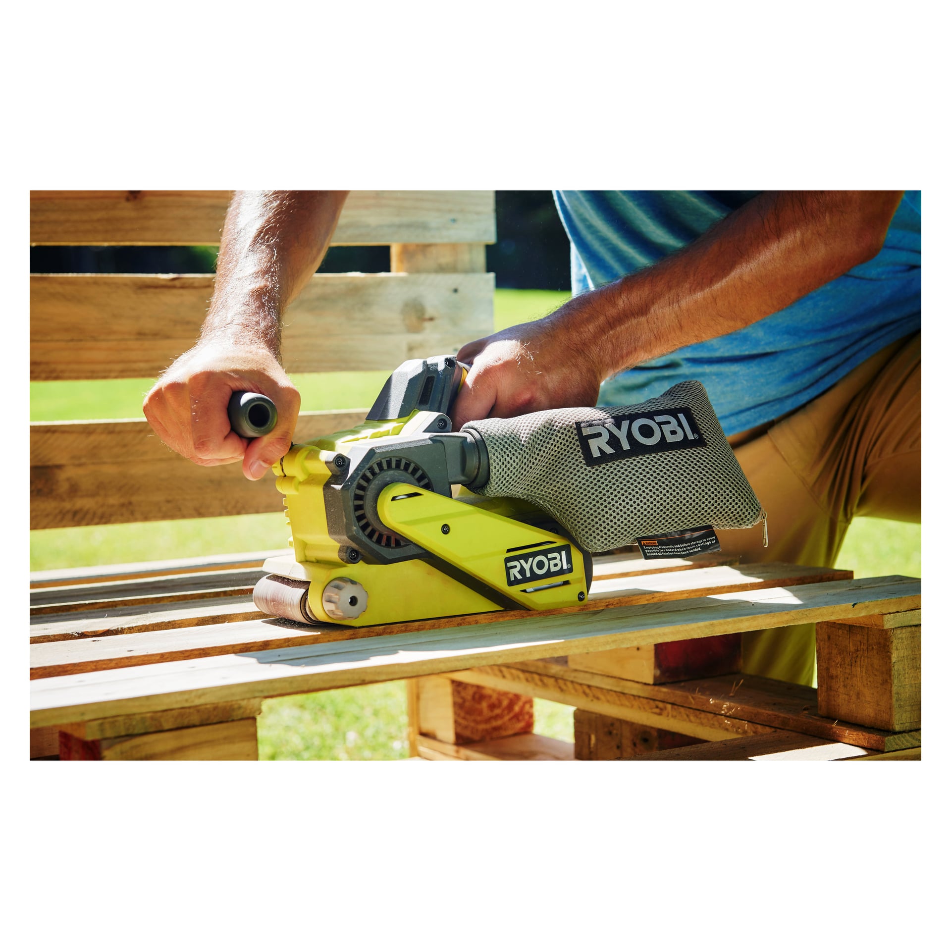 Noen bruker en gul Ryobi One+ beltsliper R18BS-0 for å pusse ned trepaller utenfor. Støv samles i stoffposen som er festet til verktøyet. Sollyset lyser opp området, og det er grønt gress i bakgrunnen.