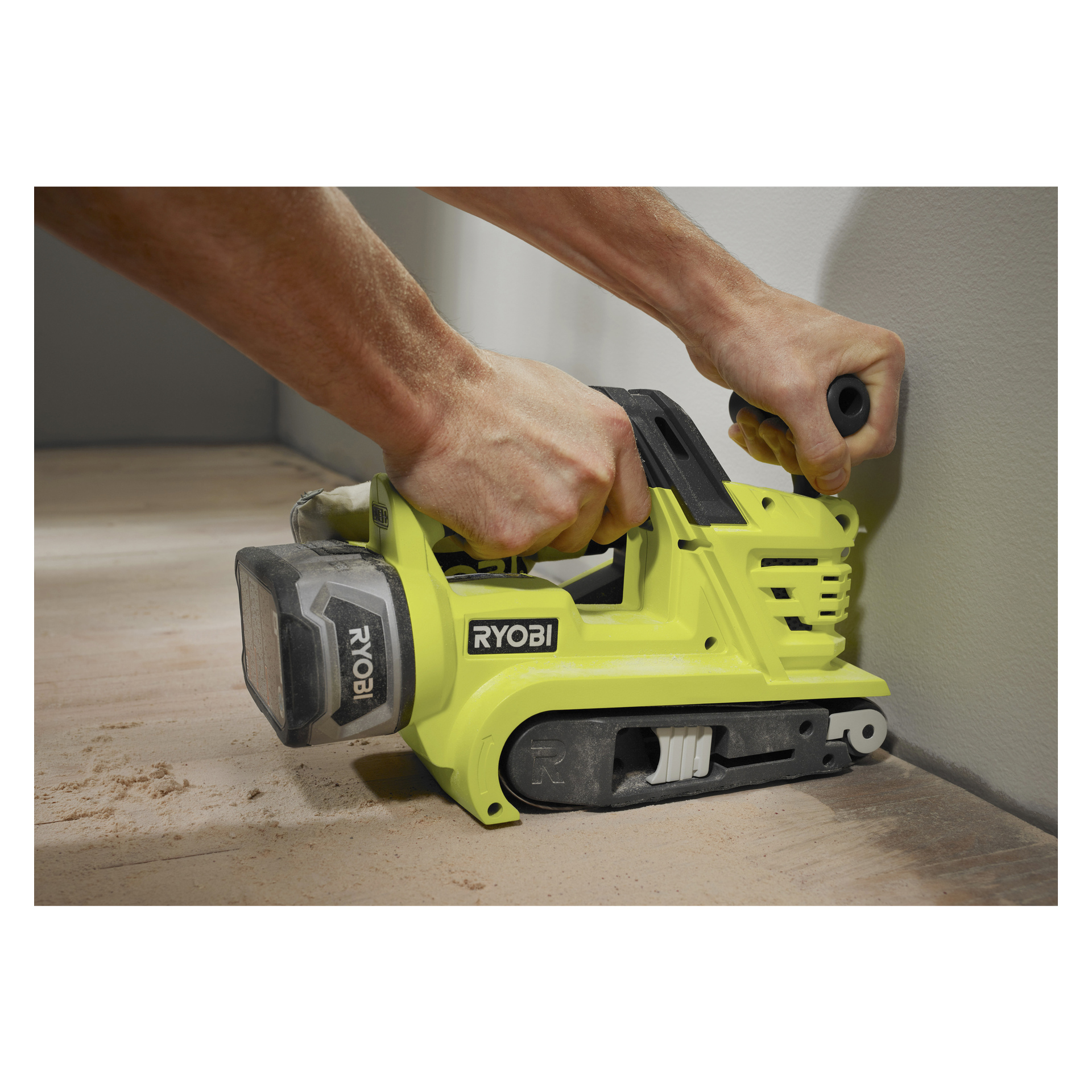 En person bruker en gul Ryobi One+ Belt-sliper R18BS-0, og sliper et tregulv nær veggen, med synlig sagflis spredt utover gulvet.