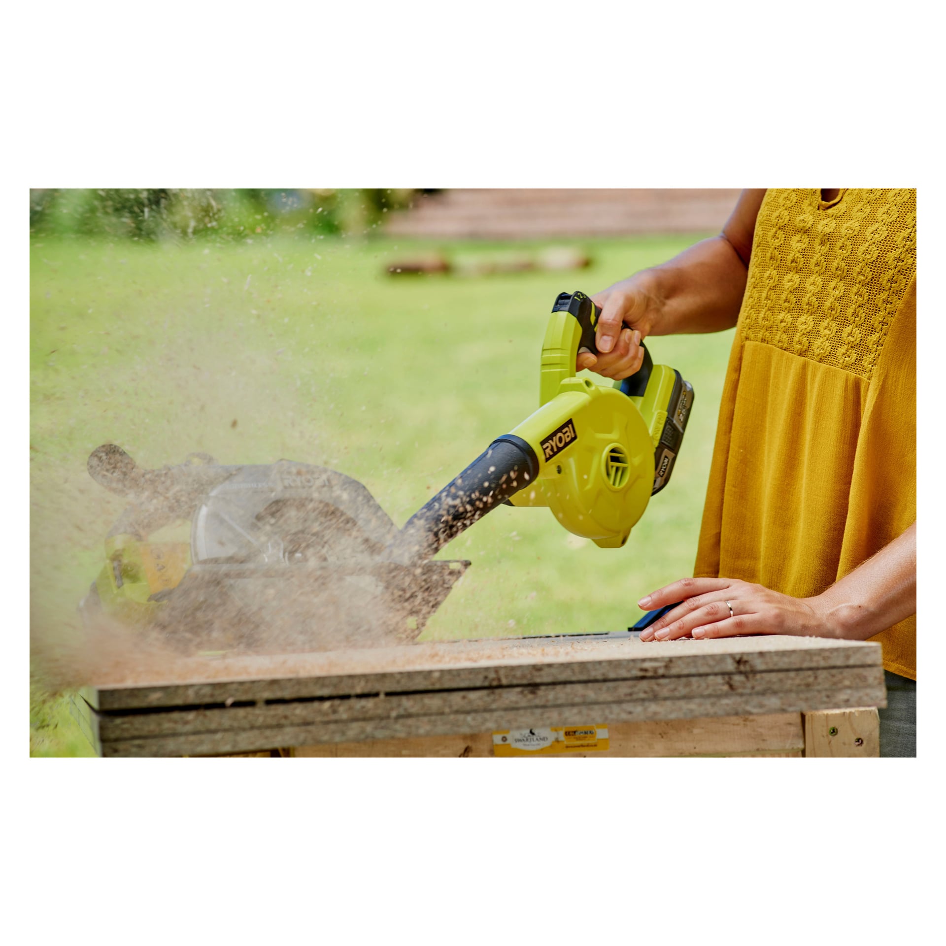 Ktoś ma na sobie żółtą bluzkę, używając zielonego Ryobi One+ Blower R18TB-0, by zdmuchnąć trociny z zewnętrznego drewnianego stołu warsztatowego. Twarz osoby jest odwrócona, a trociny wyraźnie unoszą się w powietrzu.