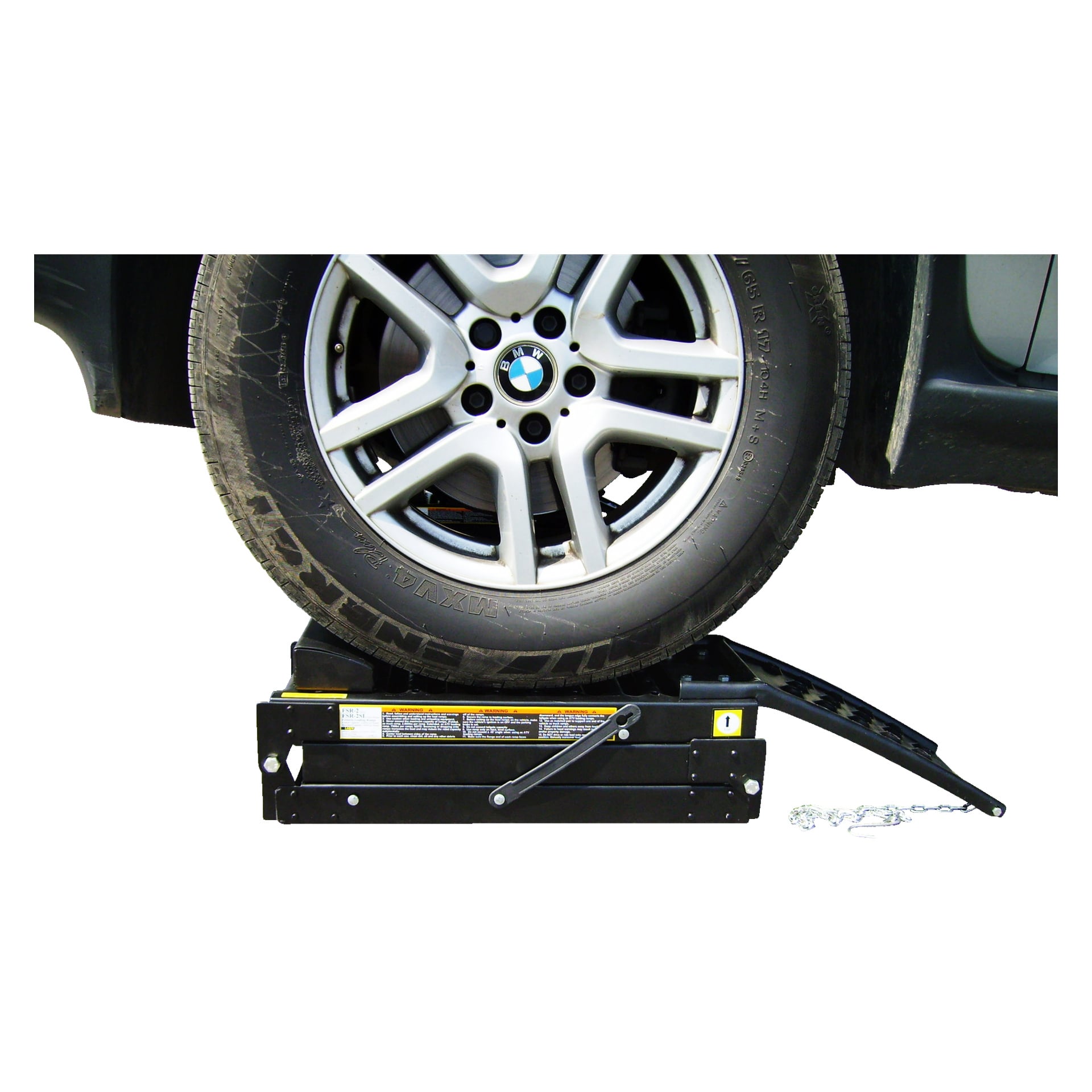 Szczegółowe zbliżenie opony BMW ustawionej na Hamron Load Ramp 2 Pack, z widocznymi fragmentami samochodu i nawierzchni. Rampa wyposażona jest w ręczną korbę oraz widoczne etykiety ostrzegawcze.