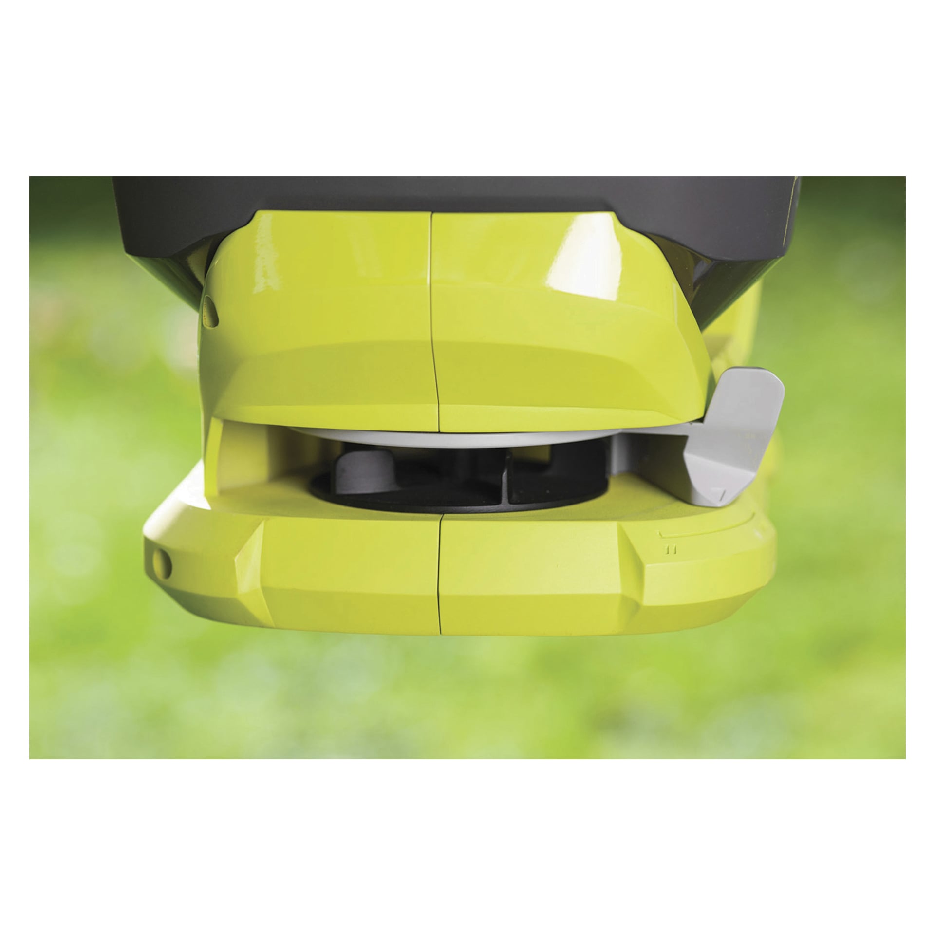 Detaljerad närbild av undersidan av Ryobi One+ OSS1800 batteridriven spridare, som visar dess livfulla gula och grå färgschema, synliga spridningsmekanism och skyddande hölje, med en mjukfokuserad grön gräsbakgrund.