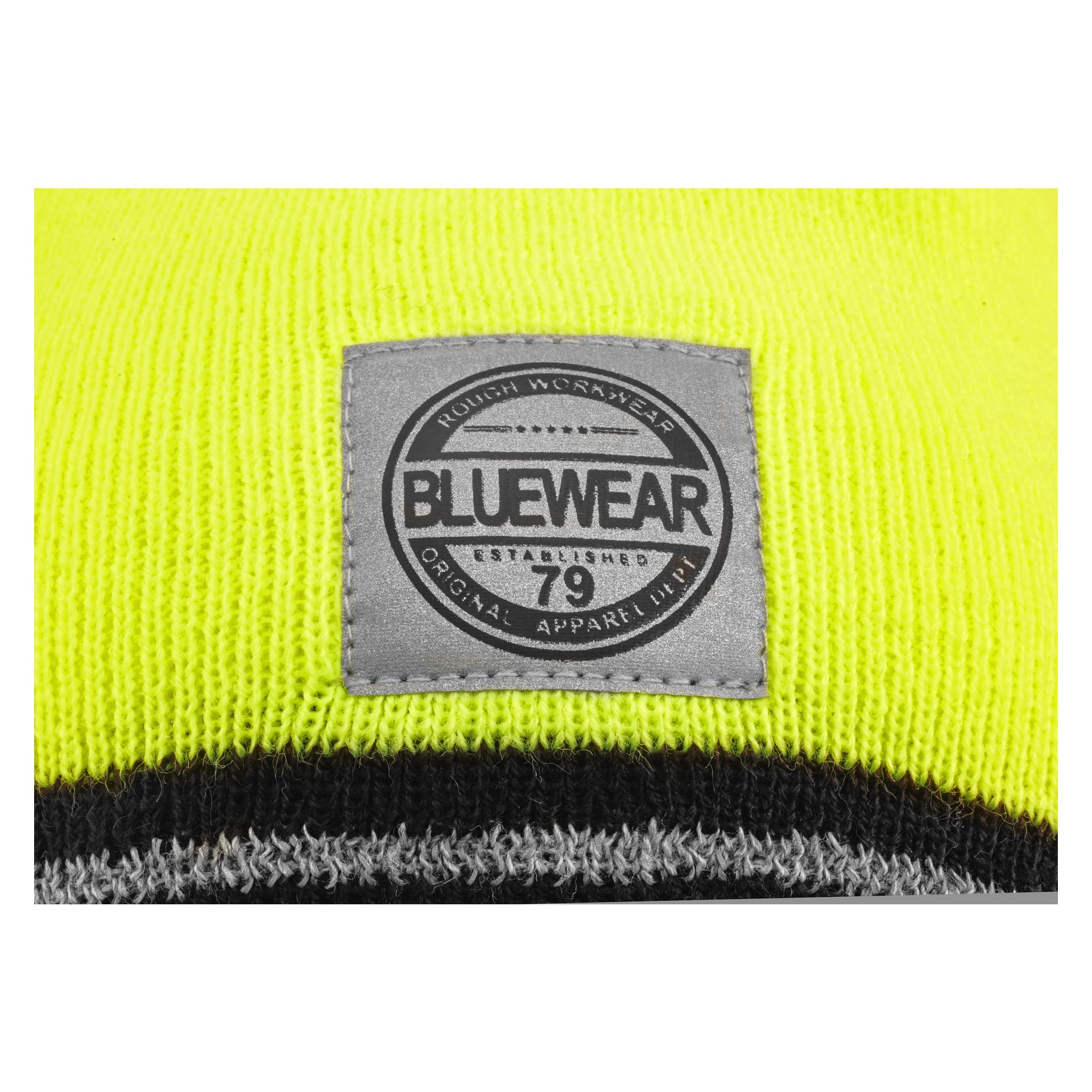 Ein graues, rechteckiges Stoffetikett wird auf gelb-schwarzes Strickmaterial genäht, Teil einer Bluewear-Mütze. Das Etikett zeigt den Markennamen 'BLUEWEAR' in großen Buchstaben sowie die Phrasen 'ROUGH WORKWEAR', 'ESTABLISHED 79' und 'ORIGINAL APPAREL LINE'.