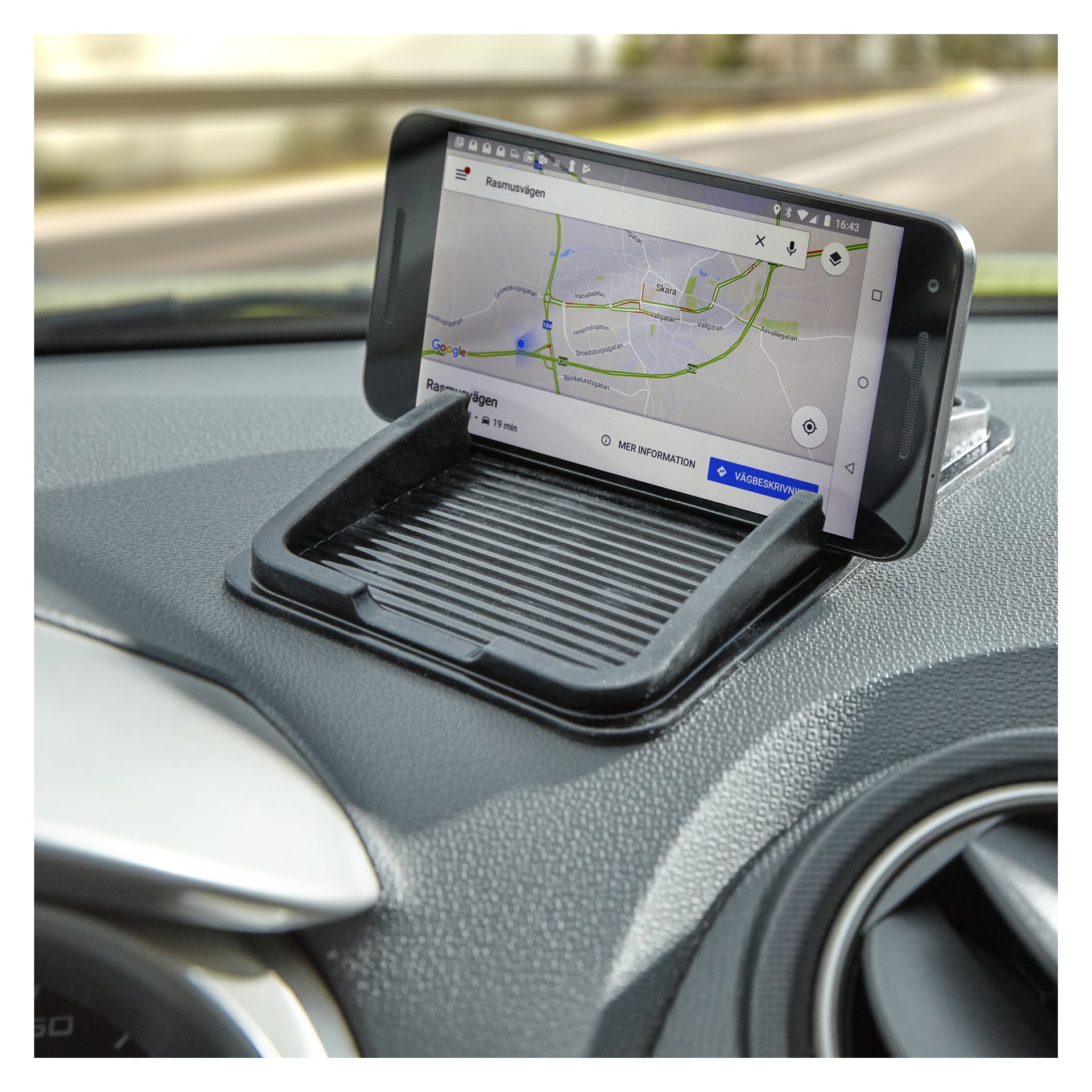 En Marquant Universal-hållare håller en smartphone i liggande läge på en bils instrumentpanel bredvid en luftventil. Telefonskärmen visar en navigationskarta, och vägen kan ses genom vindrutan framför.