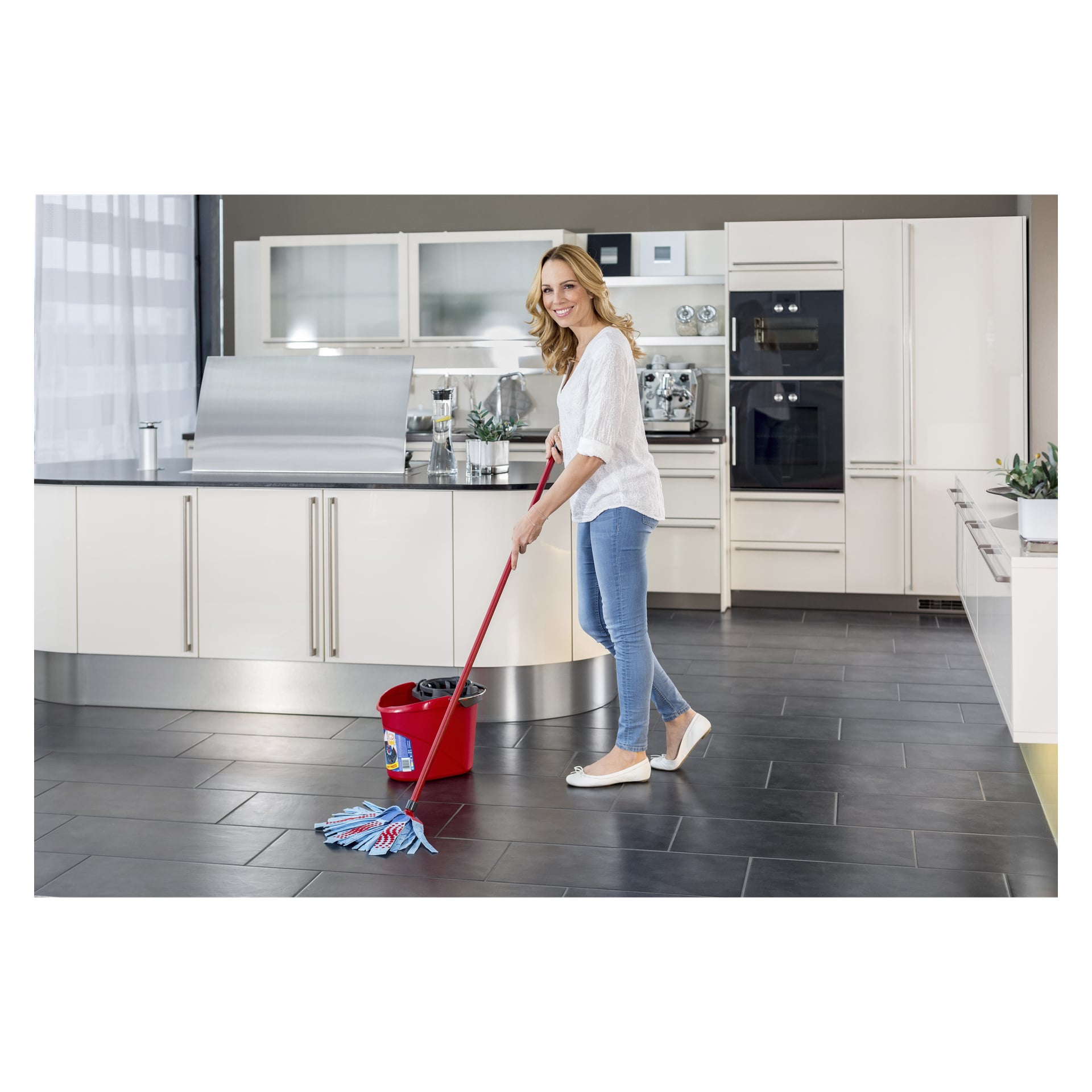 En kvinna klädd avslappnat moppar golvet i ett ljust och modernt kök. Hon använder en Vileda SuperMocio 3 Action mopp och åtföljs av en röd Vileda-mopphink med squeezer. Köket har vita skåp och rostfria apparater.