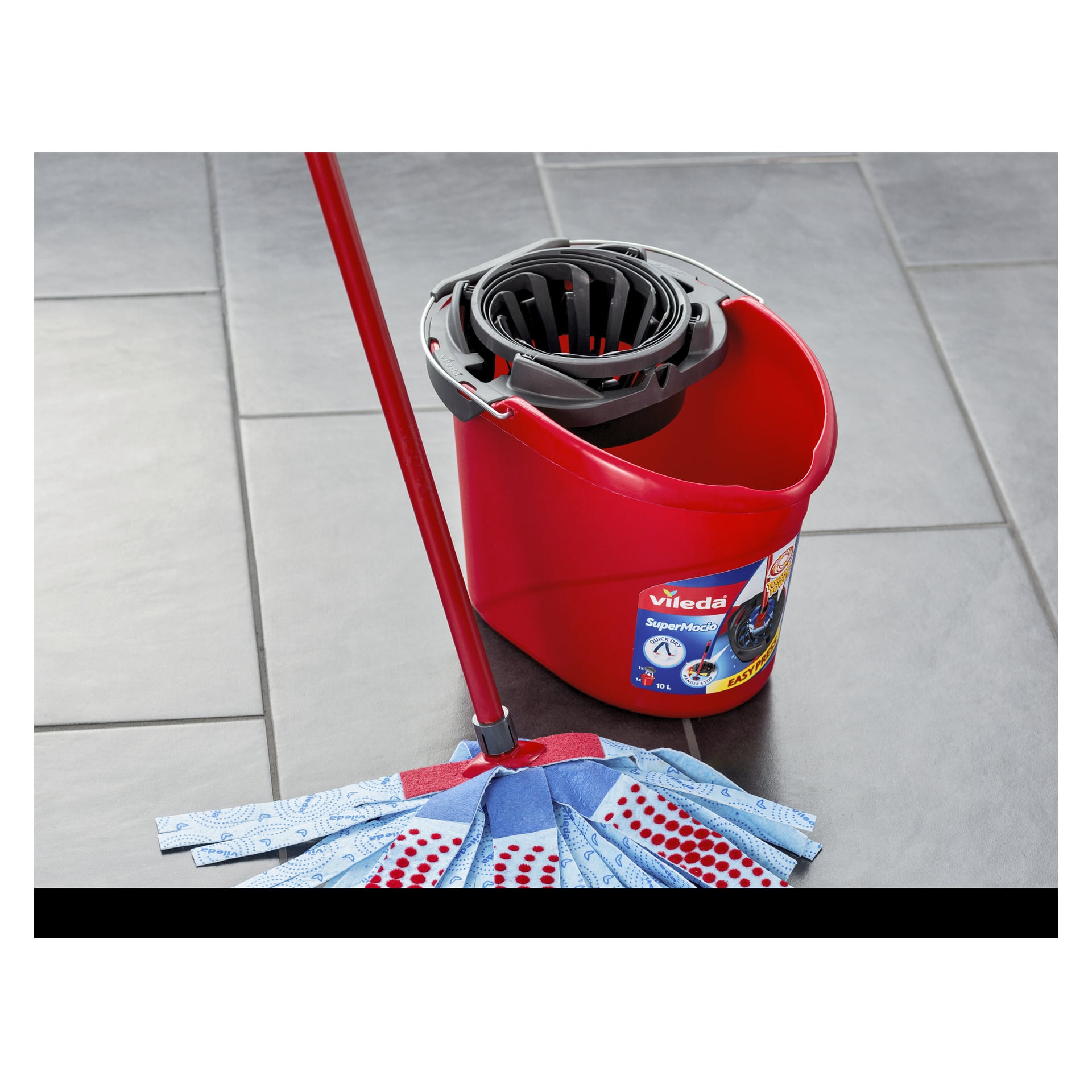 Ett Vileda Mop SuperMocio 3 Action-set, bestående av en röd hink med en wringer och en blåvit mopp med mönstrade färg, står på ett grått kaklat golv. Sidan av hinken visar en tydlig produktinformationsetikett.
