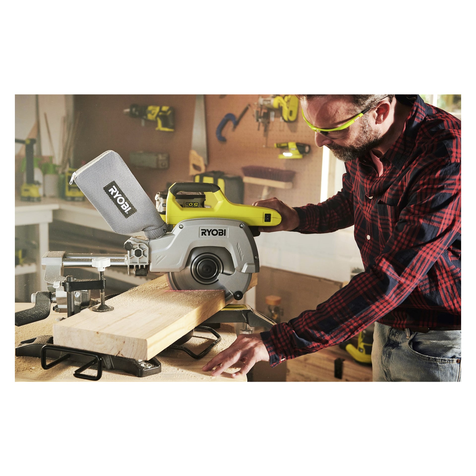 Mies turvalaseissa käyttää Ryobi One+ -paristokäyttöistä jiirisahaa (R18MS216-0) leikatakseen puulaudan työpajassa, jota ympäröivät työkalut ja laitteet, jotka on asetettu hyllyille ja seinälle hänen takanaan.